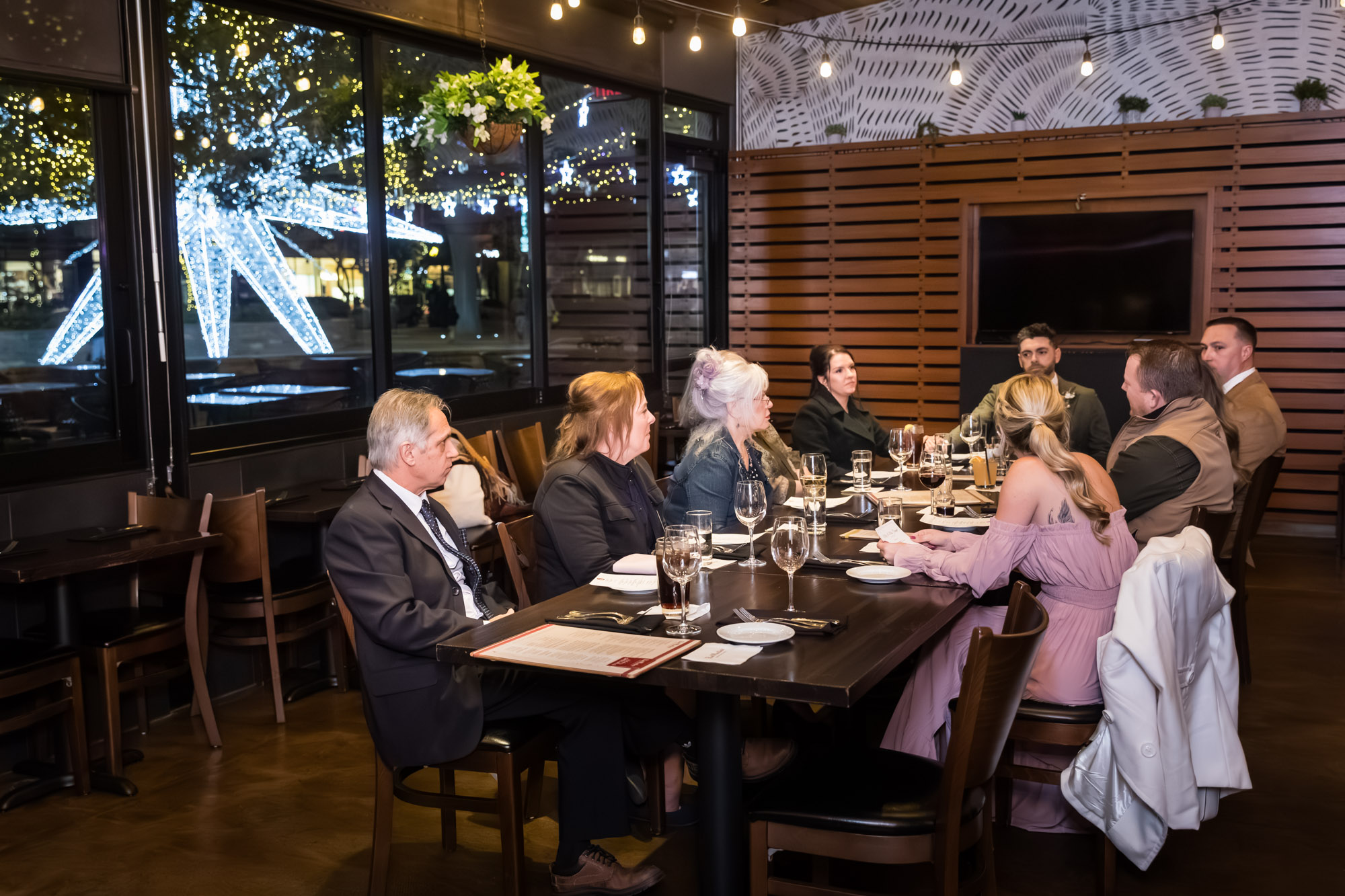 Guests seated at long table in private room against wall with windows at a wedding reception