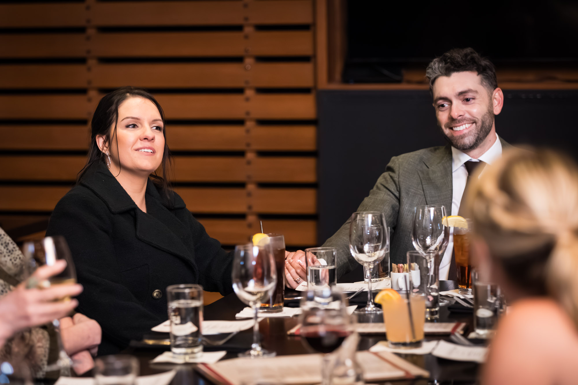 Man wearing grey suit jacket seated next to woman wearing black jacket in wood-paneled room during wedding reception