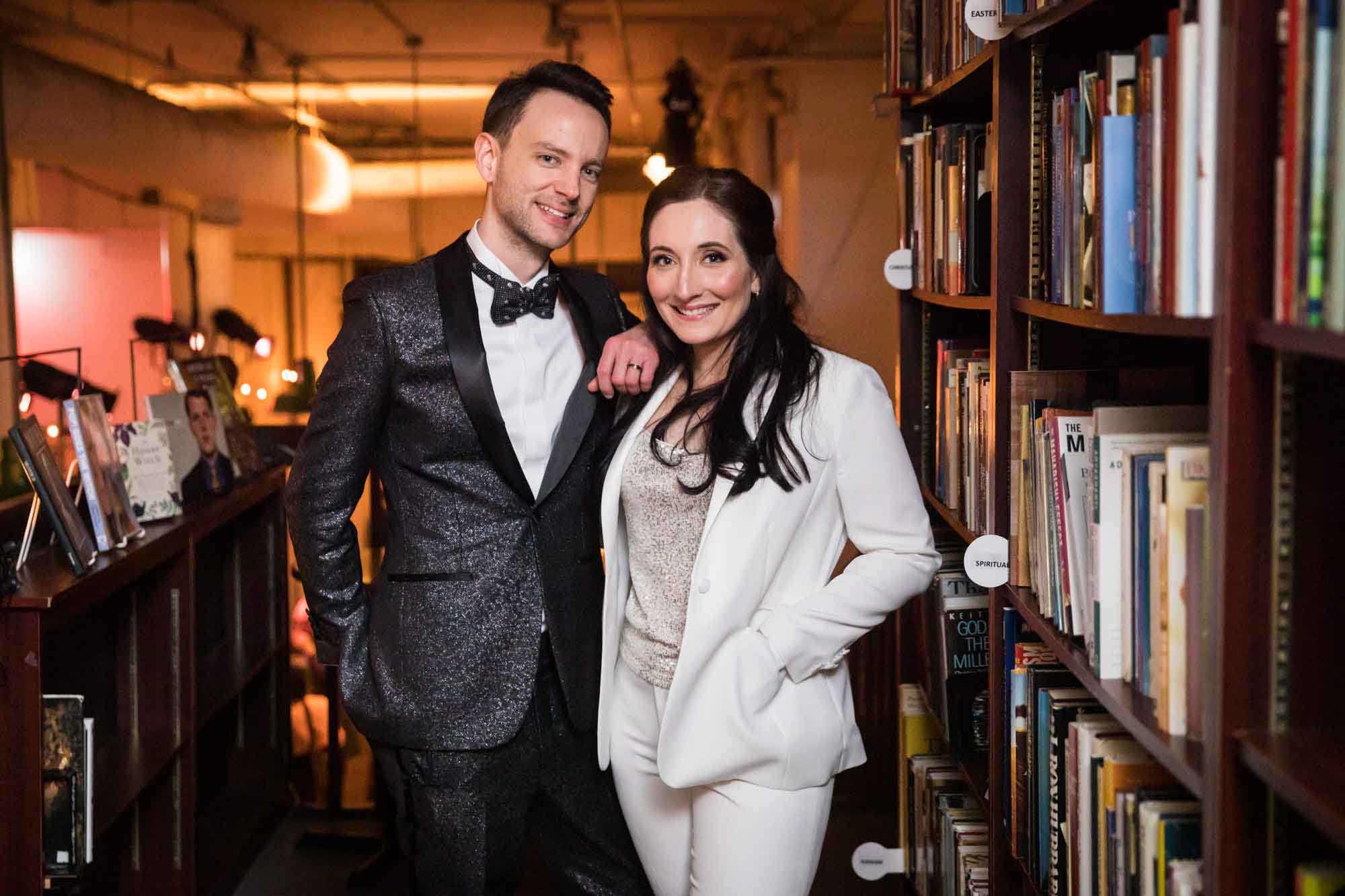 Bride wearing white suit standing with groom wearing black shiny suit in between bookcases for article on how to elope in San Antonio