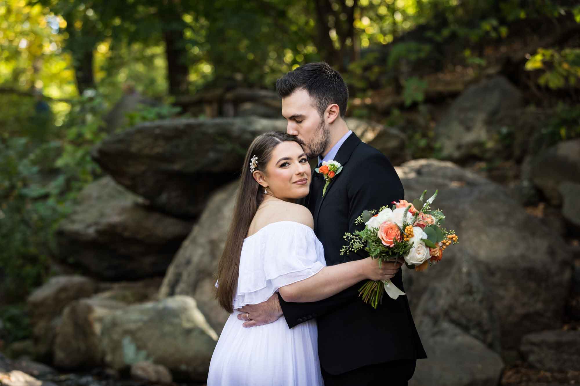 Bride wearing off-shoulder white dress holding flower bouquet being kissed on head by groom in front of rocks and trees for article on how to elope in San Antonio