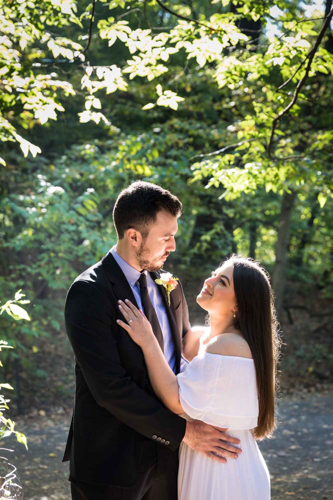 Bride wearing off-shoulder white dress looking up at groom wearing black suit in front of trees for article on how to elope in San Antonio