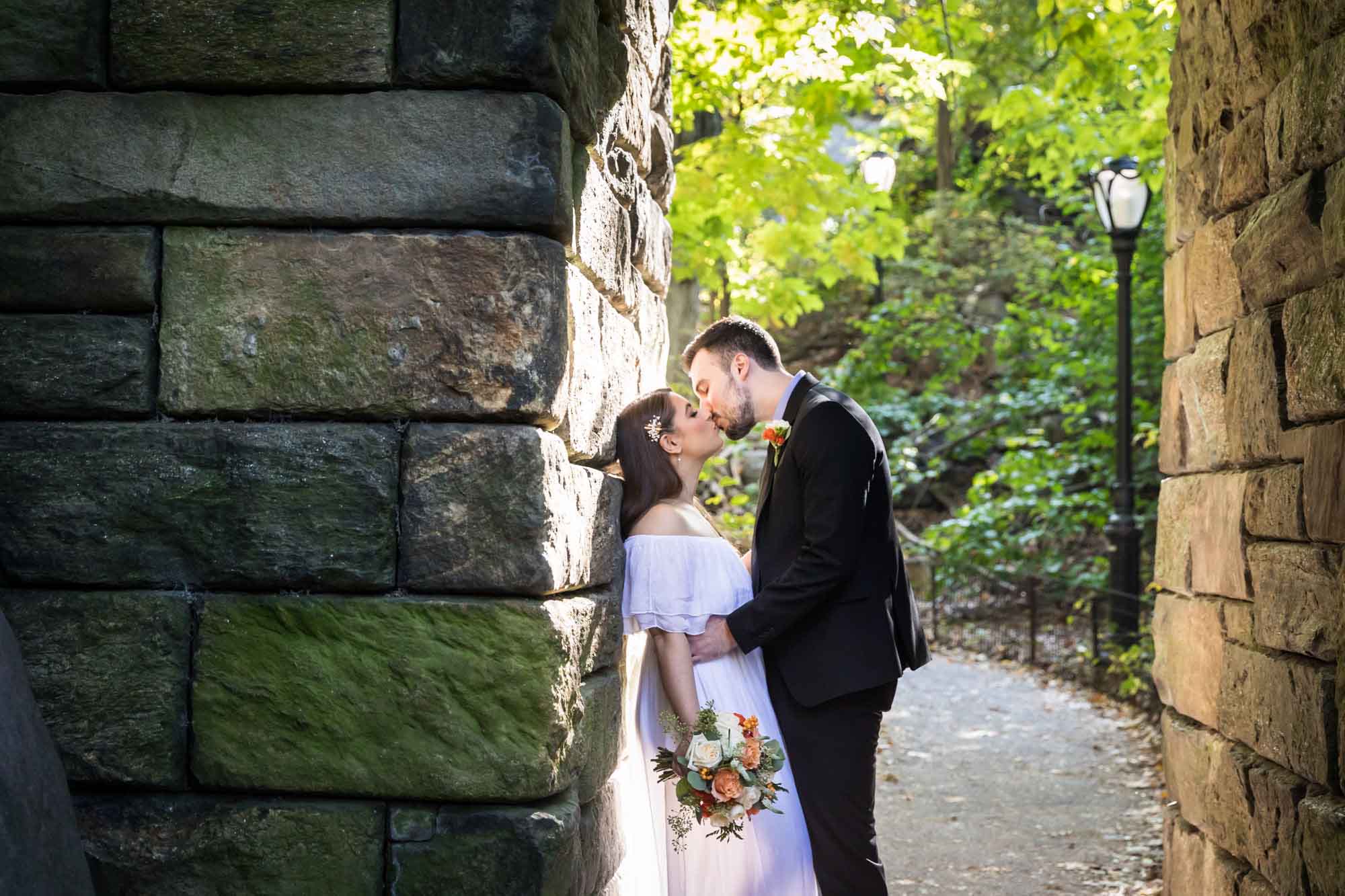 Bride wearing off-shoulder white dress kissing groom wearing black suit against stone wall in park for article on how to elope in San Antonio