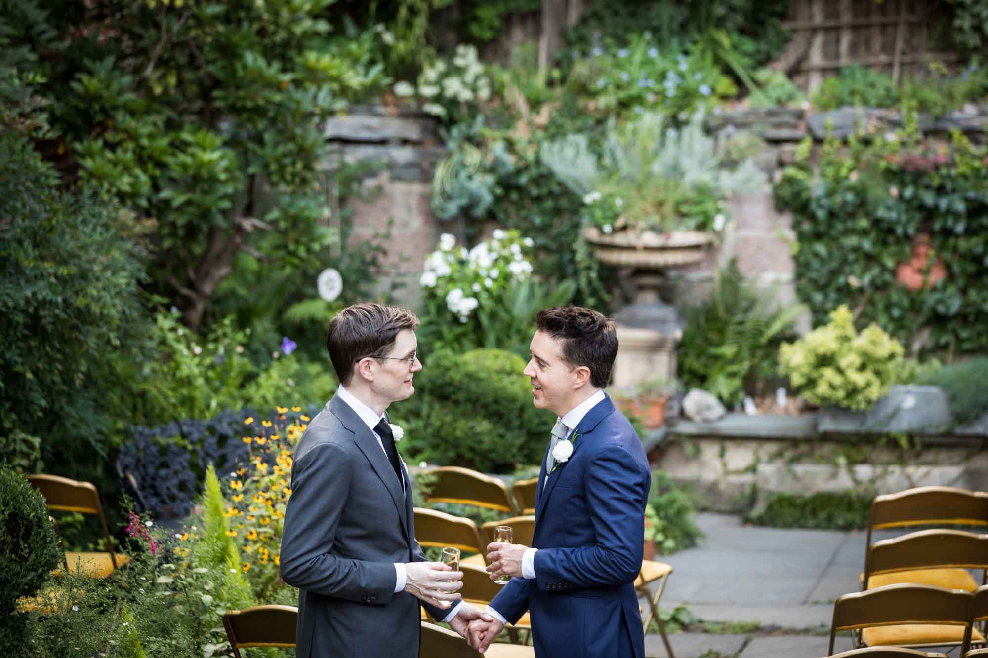 Two grooms holding hands and holding champagne glasses in a park filled with flowers for article on how to elope in San Antonio