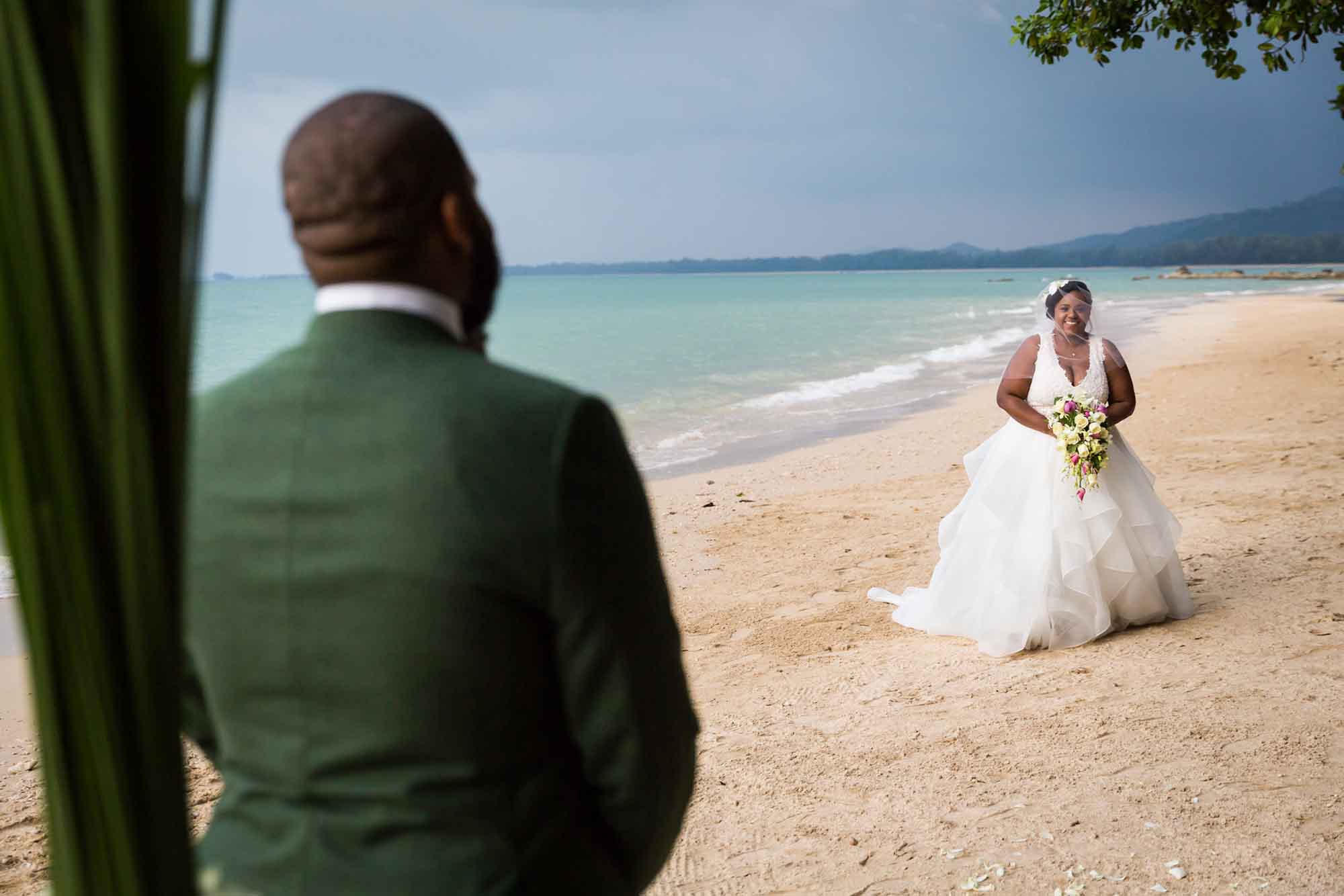 Bride wearing white dress holding flower bouquet walking down beach in Thailand to groom wearing green jacket for article on how to elope in San Antonio