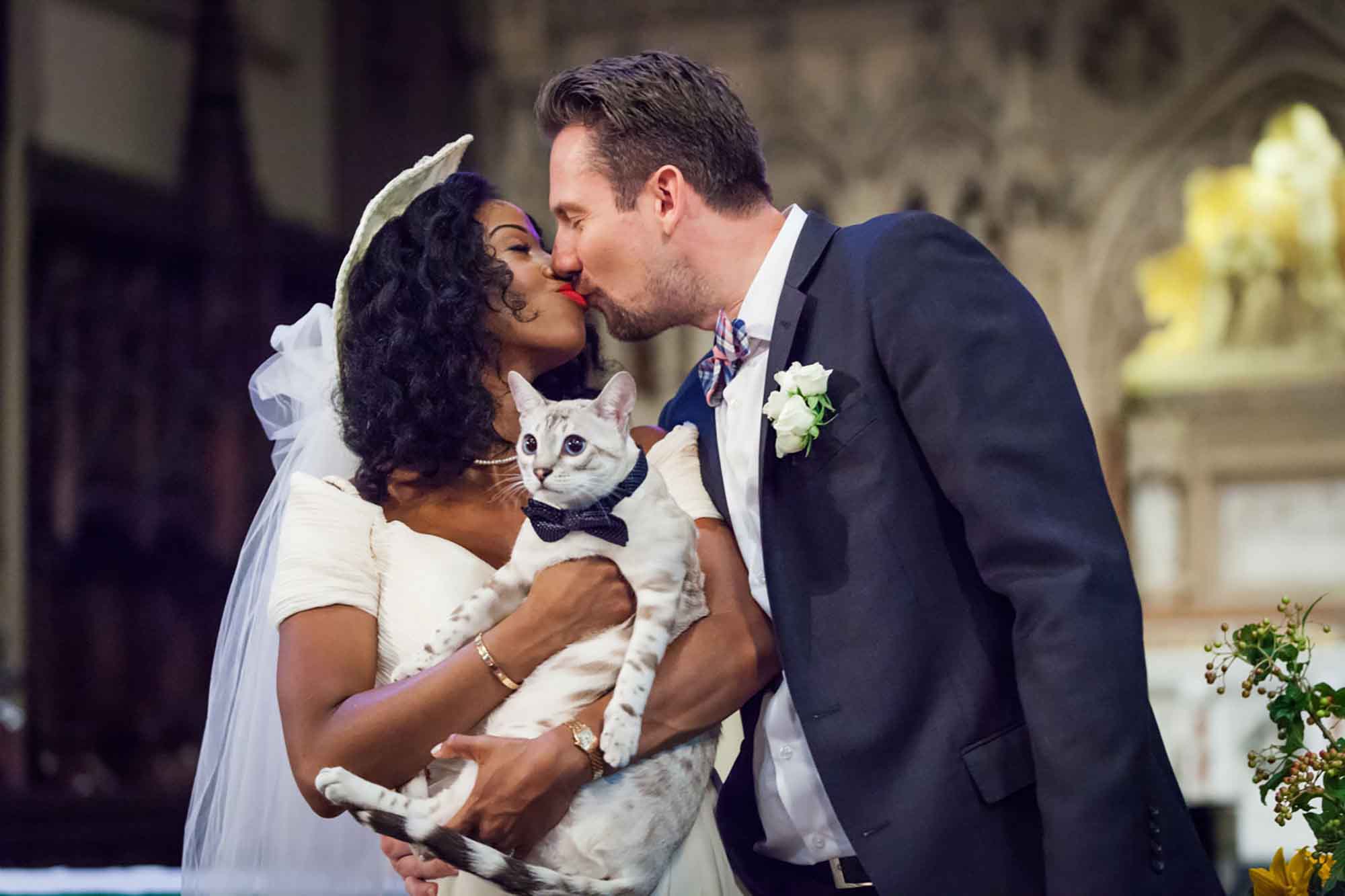 Groom kissing bride wearing hat and white veil while holding cat in church for article on how to elope in San Antonio
