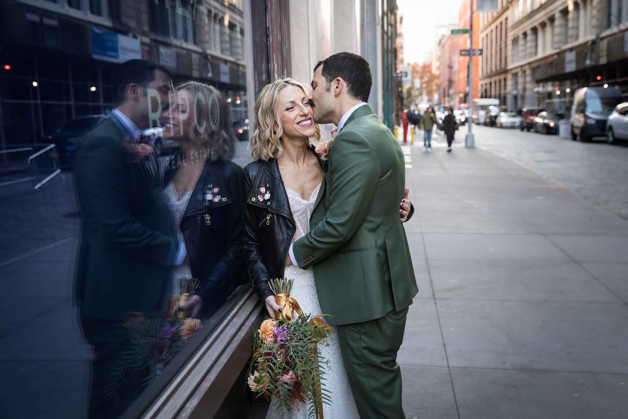 Groom wearing green suit kissing bride wearing black leather jacket up against reflective glass window on sidewalk for article on how to elope in San Antonio