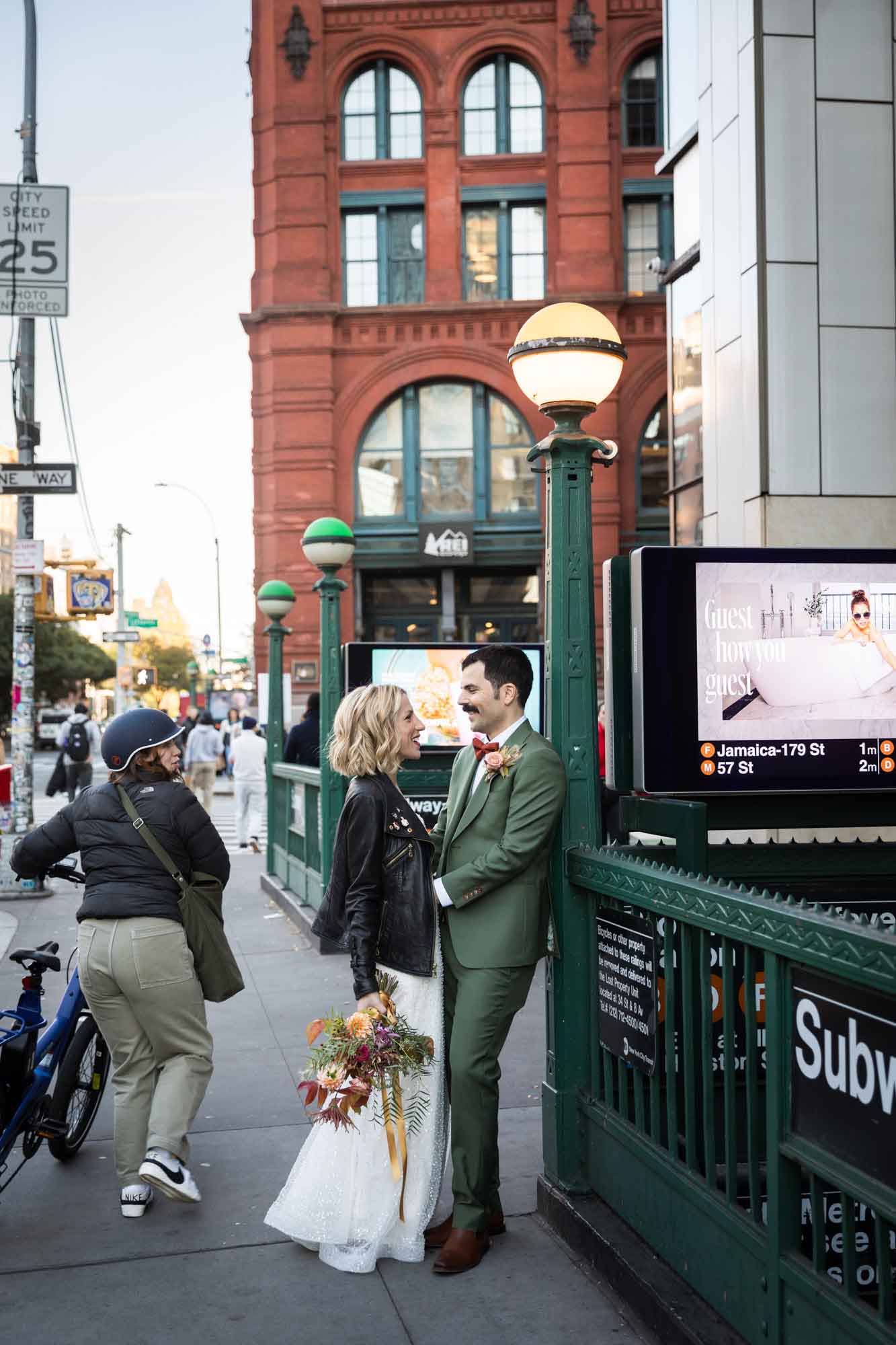Bride wearing black leather jacket standing with groom wearing green suit up against subway entrance on sidewalk in New York for article on how to elope in San Antonio