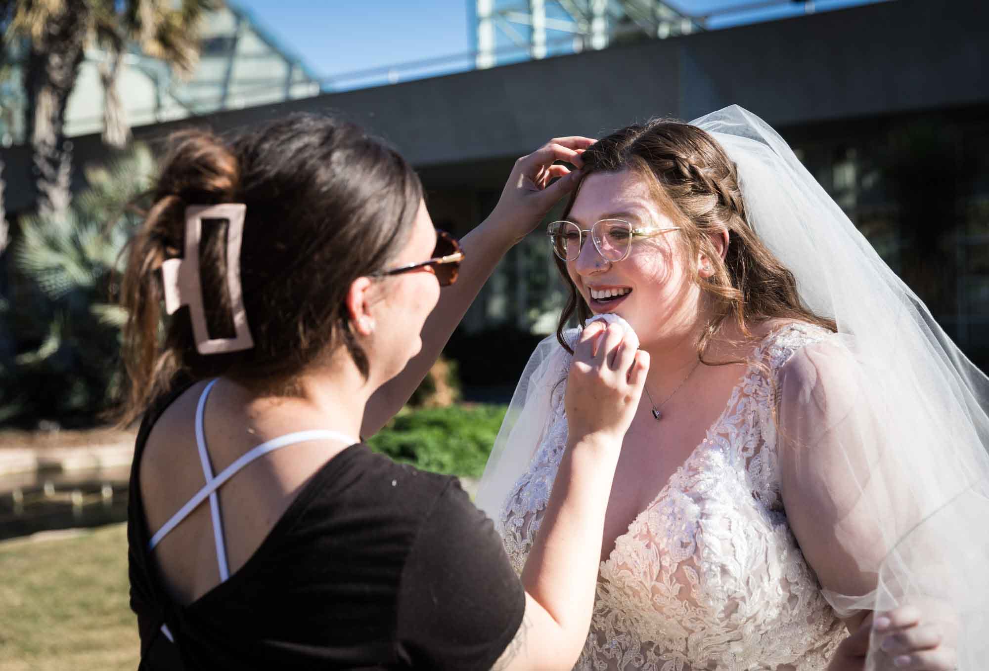 Woman with hair in clip applying lipstick to bride wearing white dress and veil during a San Antonio Botanical Gardens bridal portrait session