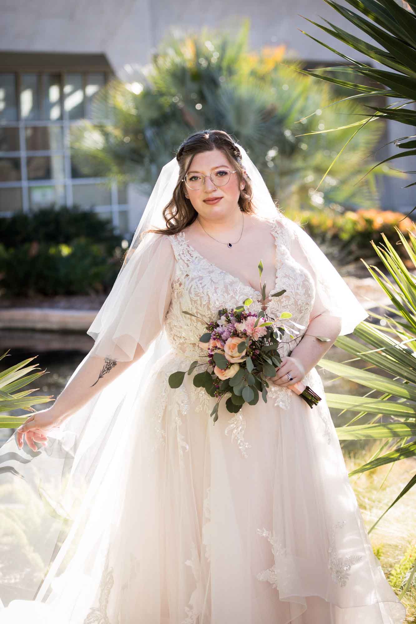 Bride wearing white dress and veil holding bouquet of flowers and hem of dress standing beside palm fronds during a San Antonio Botanical Gardens bridal portrait session