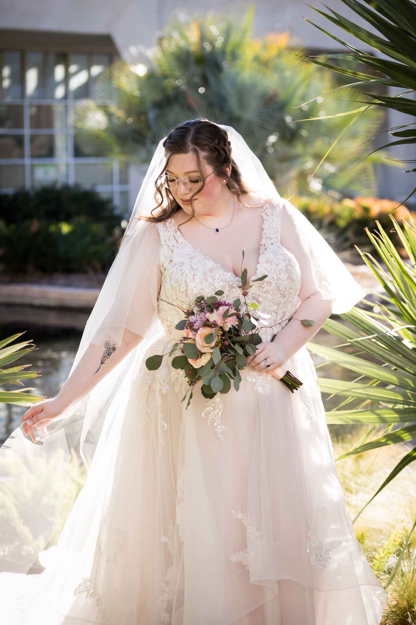 Bride wearing white dress and veil holding bouquet of flowers and hem of dress standing beside palm fronds during a San Antonio Botanical Gardens bridal portrait session