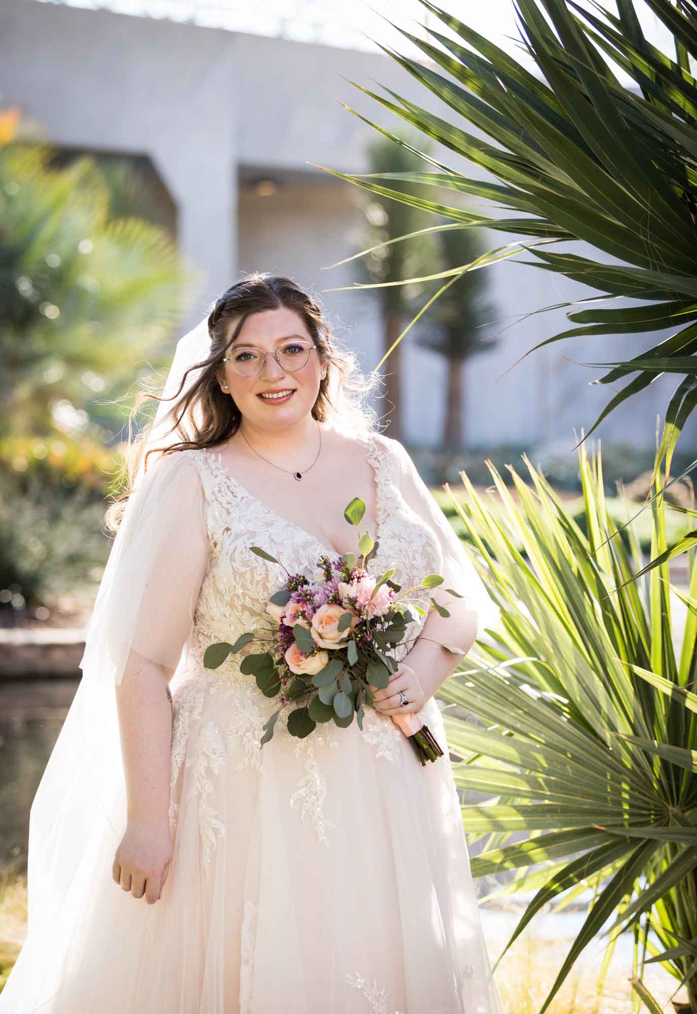 Bride wearing white dress and veil holding bouquet of flowers standing beside palm fronds during a San Antonio Botanical Gardens bridal portrait session