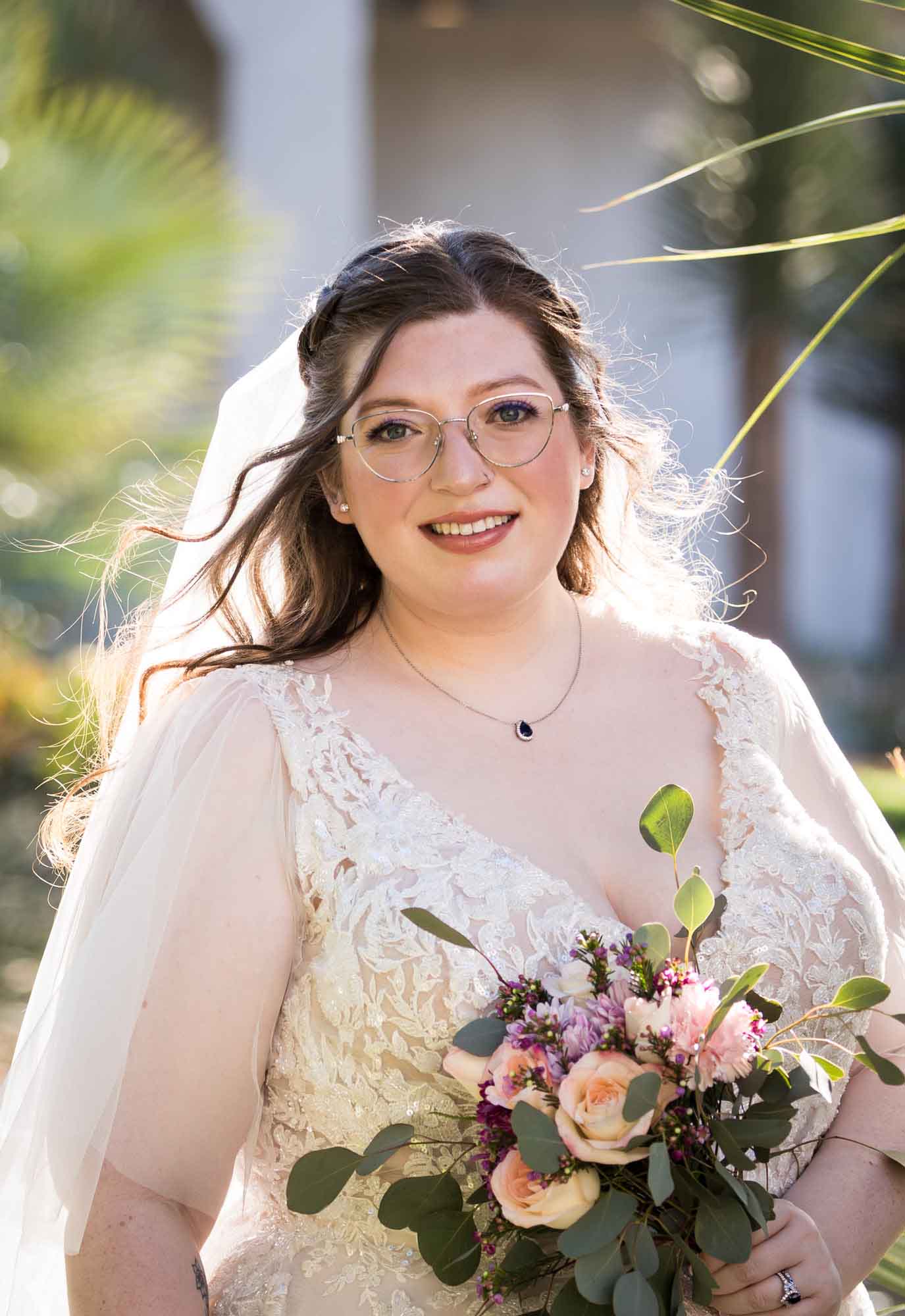 Bride wearing white dress and veil holding bouquet of flowers standing beside palm fronds during a San Antonio Botanical Gardens bridal portrait session