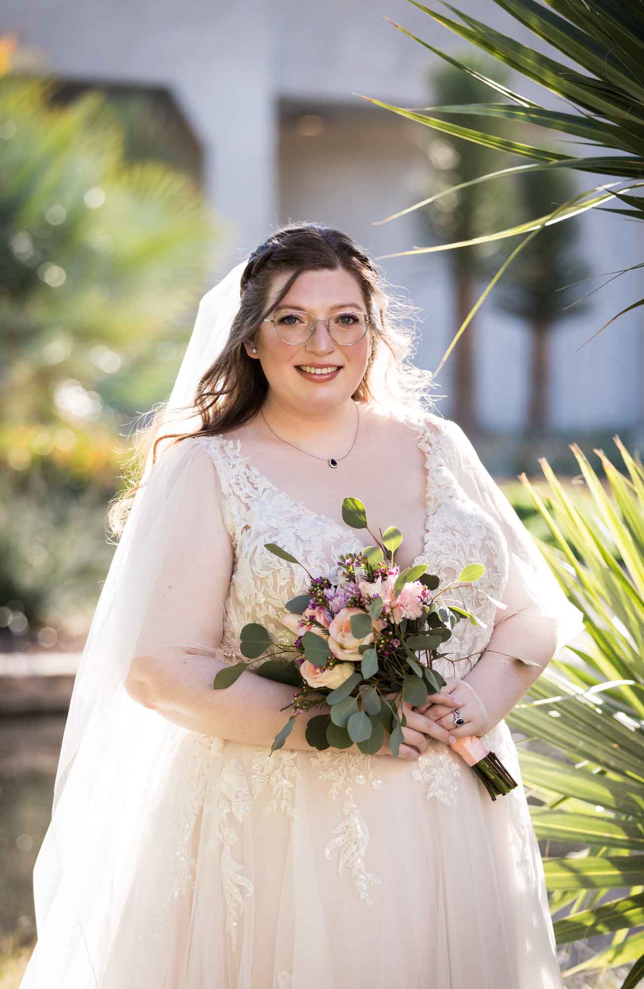 Bride wearing white dress and veil holding bouquet of flowers standing beside palm fronds during a San Antonio Botanical Gardens bridal portrait session