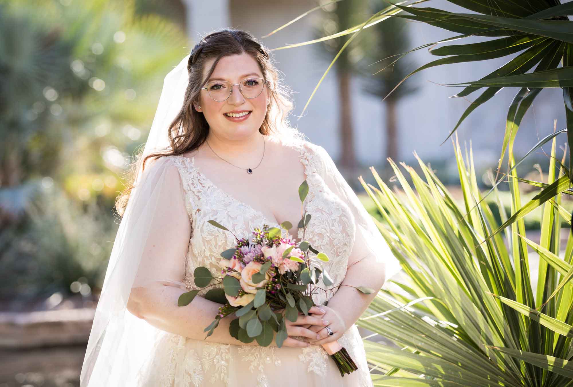 Bride wearing white dress and veil holding bouquet of flowers standing beside palm fronds during a San Antonio Botanical Gardens bridal portrait session