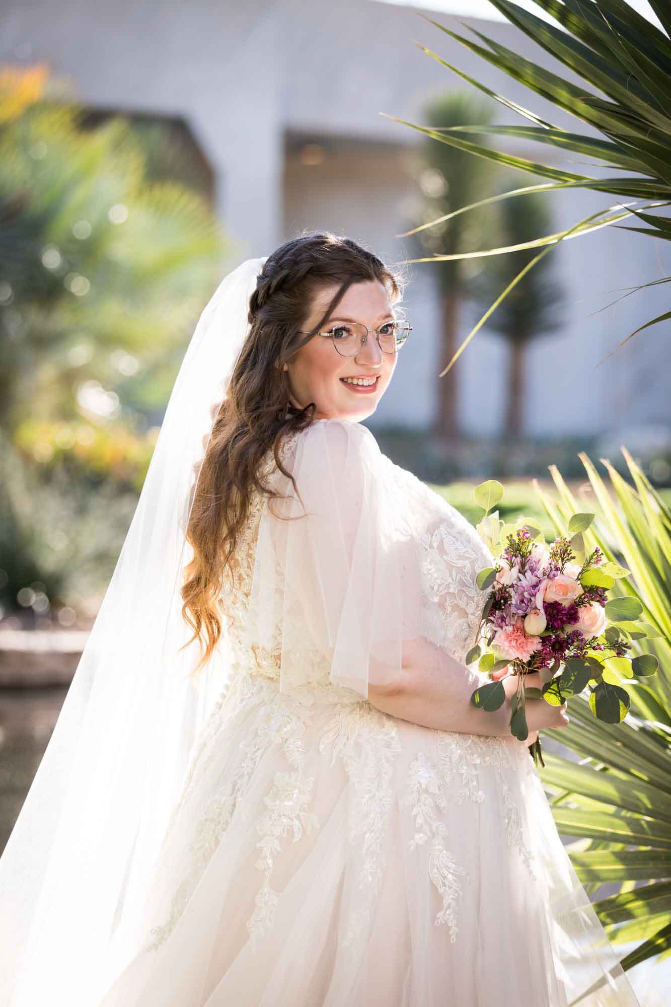 Bride wearing white dress and veil holding bouquet of flowers standing beside palm fronds during a San Antonio Botanical Gardens bridal portrait session