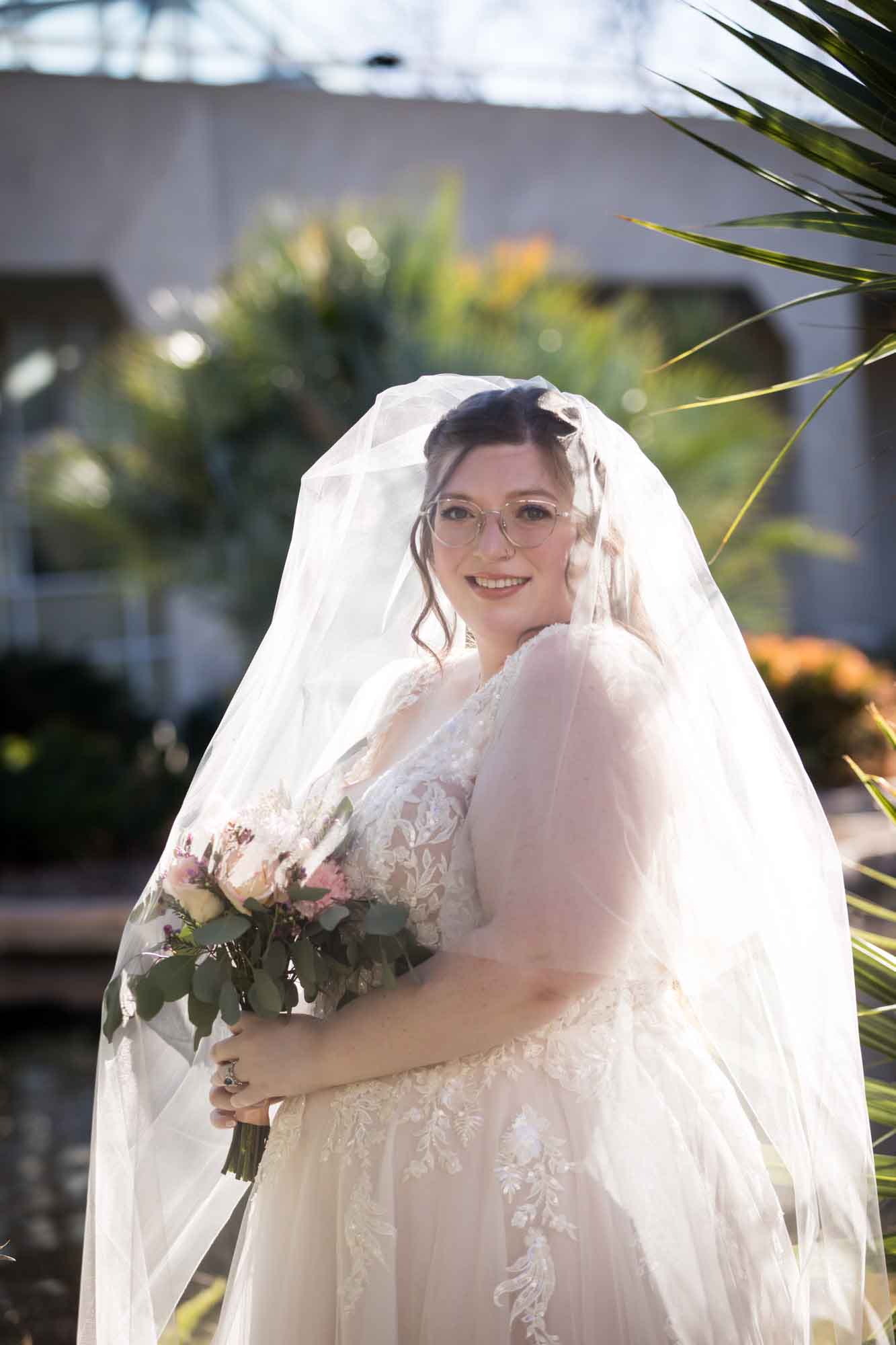 Bride wearing white dress holding bouquet of flowers with veil covering her face and body standing beside palm fronds during a San Antonio Botanical Gardens bridal portrait session