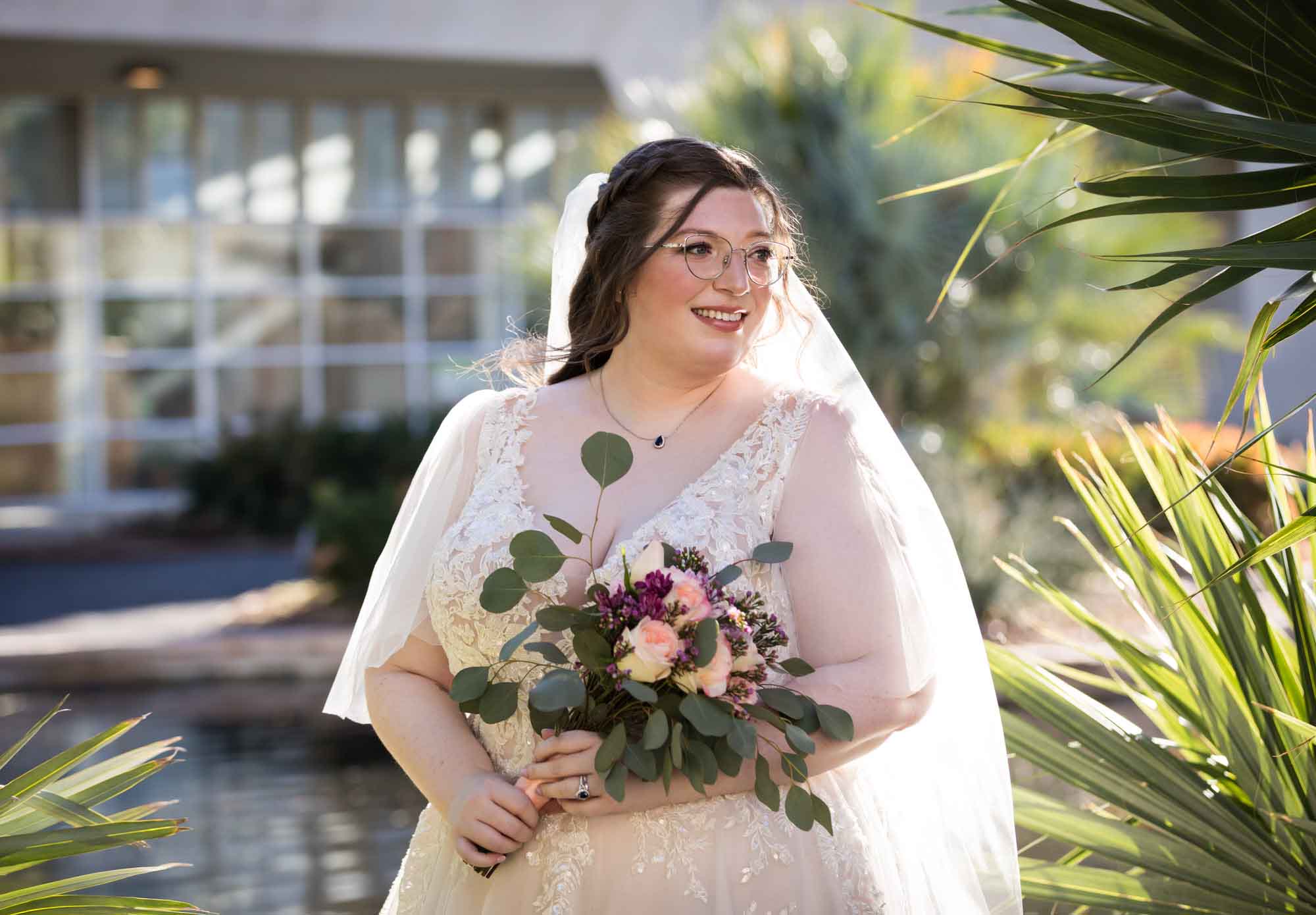 Bride wearing white dress and veil holding bouquet of flowers standing beside palm fronds during a San Antonio Botanical Gardens bridal portrait session