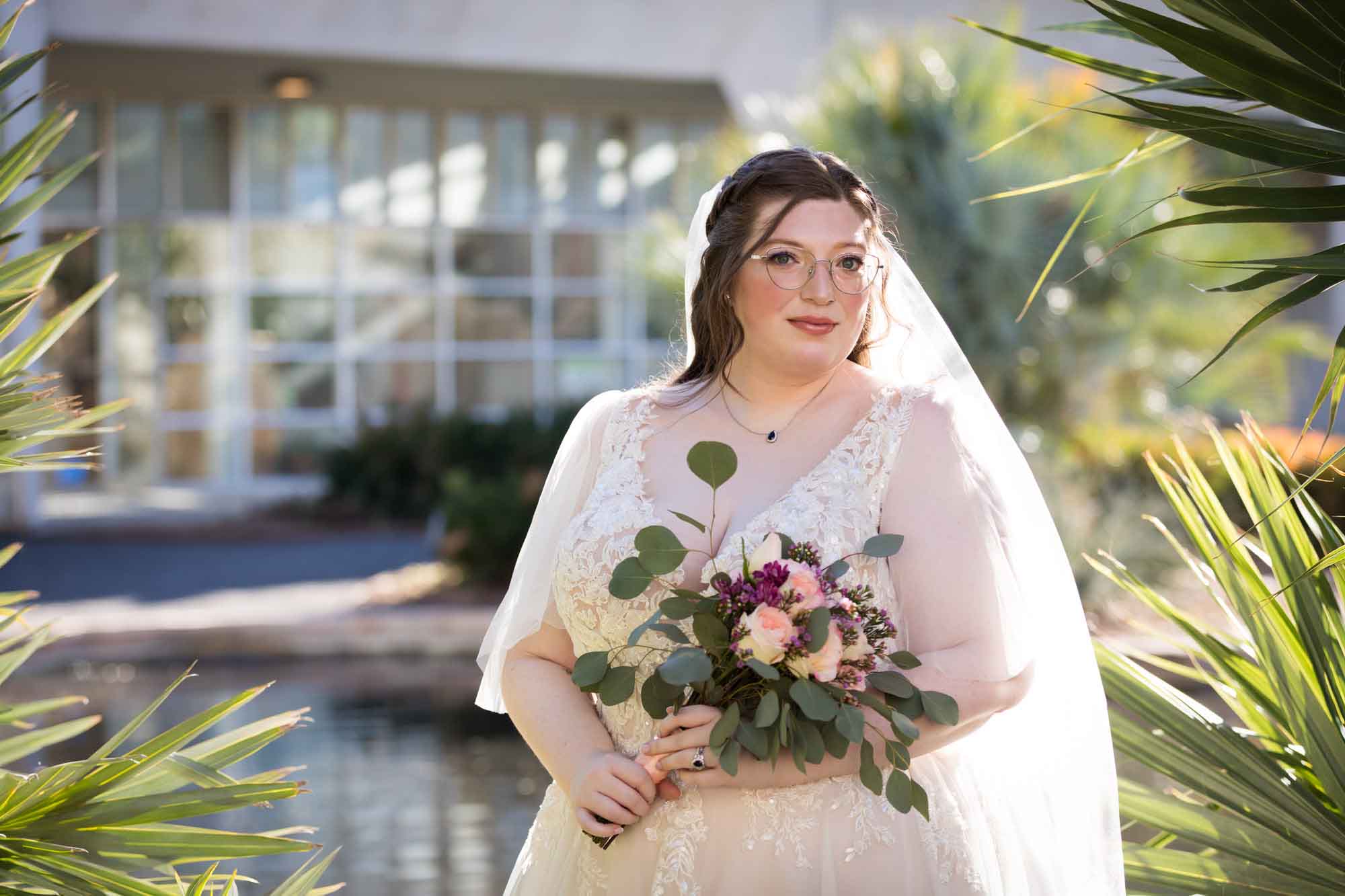 Bride wearing white dress and veil holding bouquet of flowers standing beside palm fronds during a San Antonio Botanical Gardens bridal portrait session