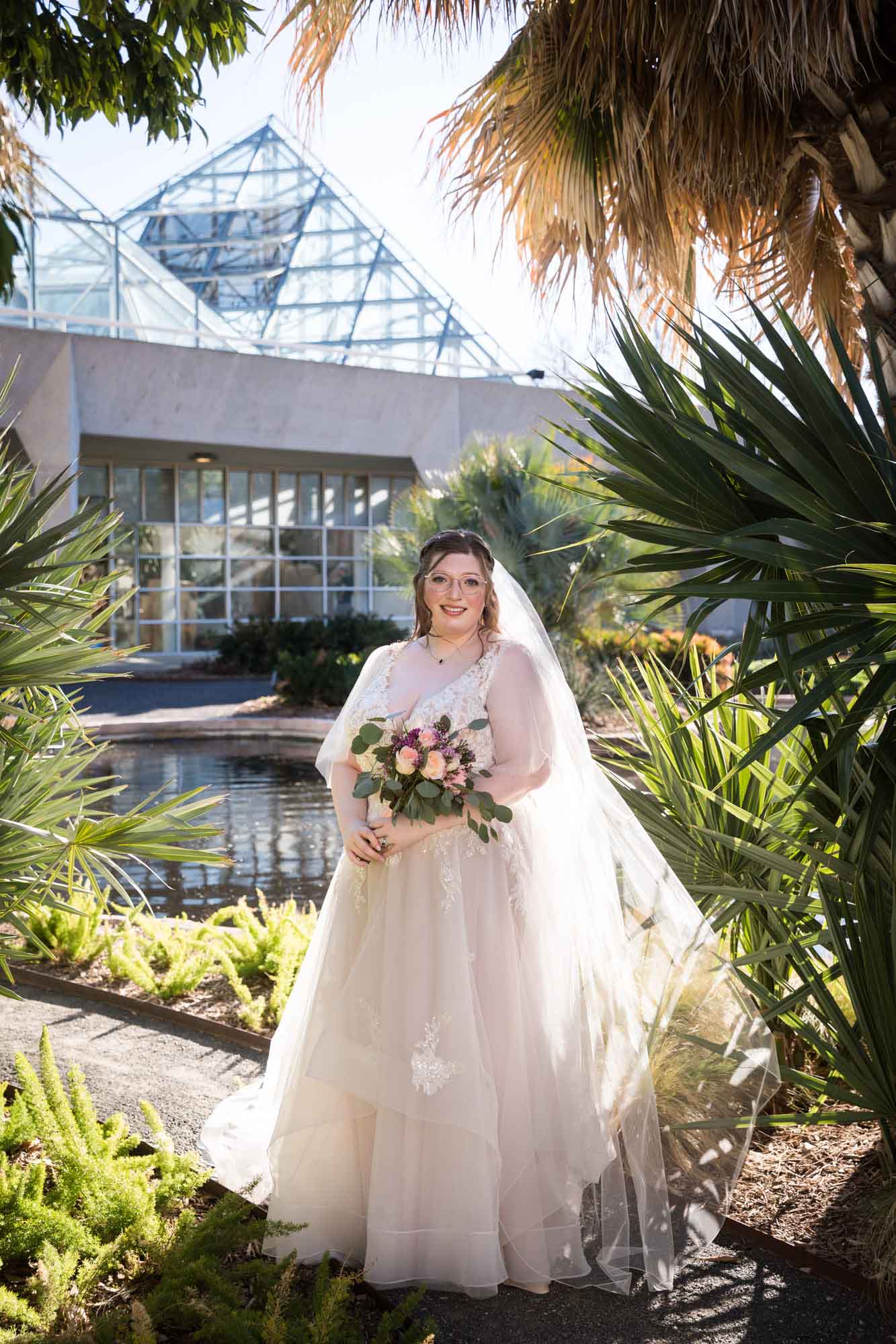 Bride wearing white dress and veil holding bouquet of flowers standing beside palm fronds during a San Antonio Botanical Gardens bridal portrait session
