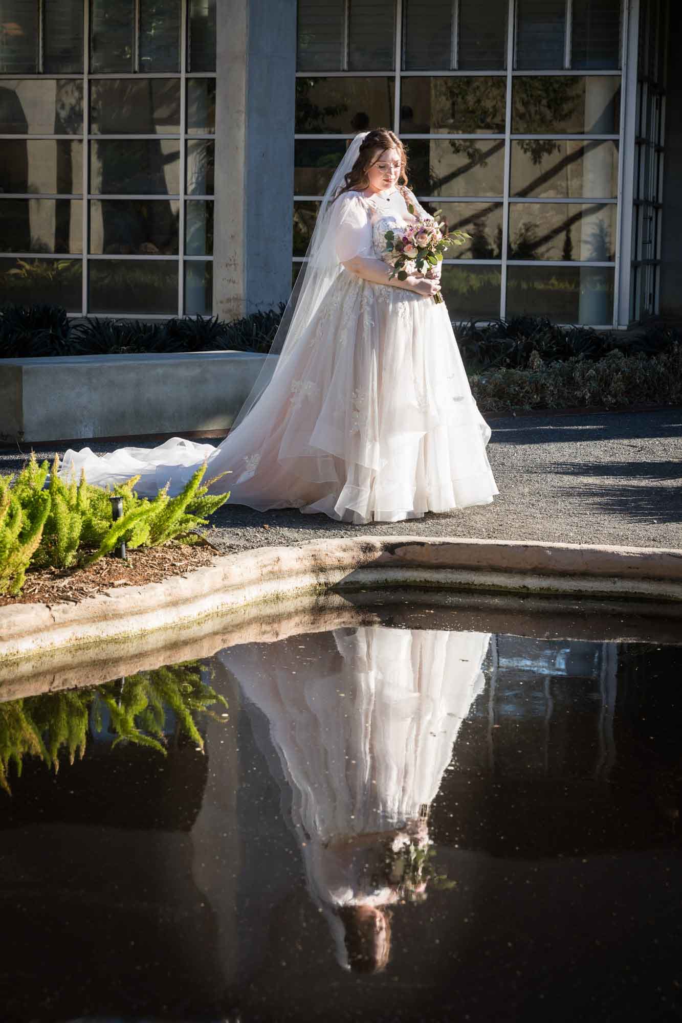 Bride wearing white dress and veil looking down at bouquet of flowers while reflection in pond during a San Antonio Botanical Gardens bridal portrait session
