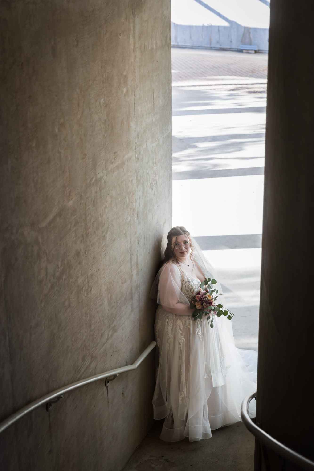 Bride wearing white dress and veil holding flower bouquet standing against concrete wall in staircase during a San Antonio Botanical Gardens bridal portrait session