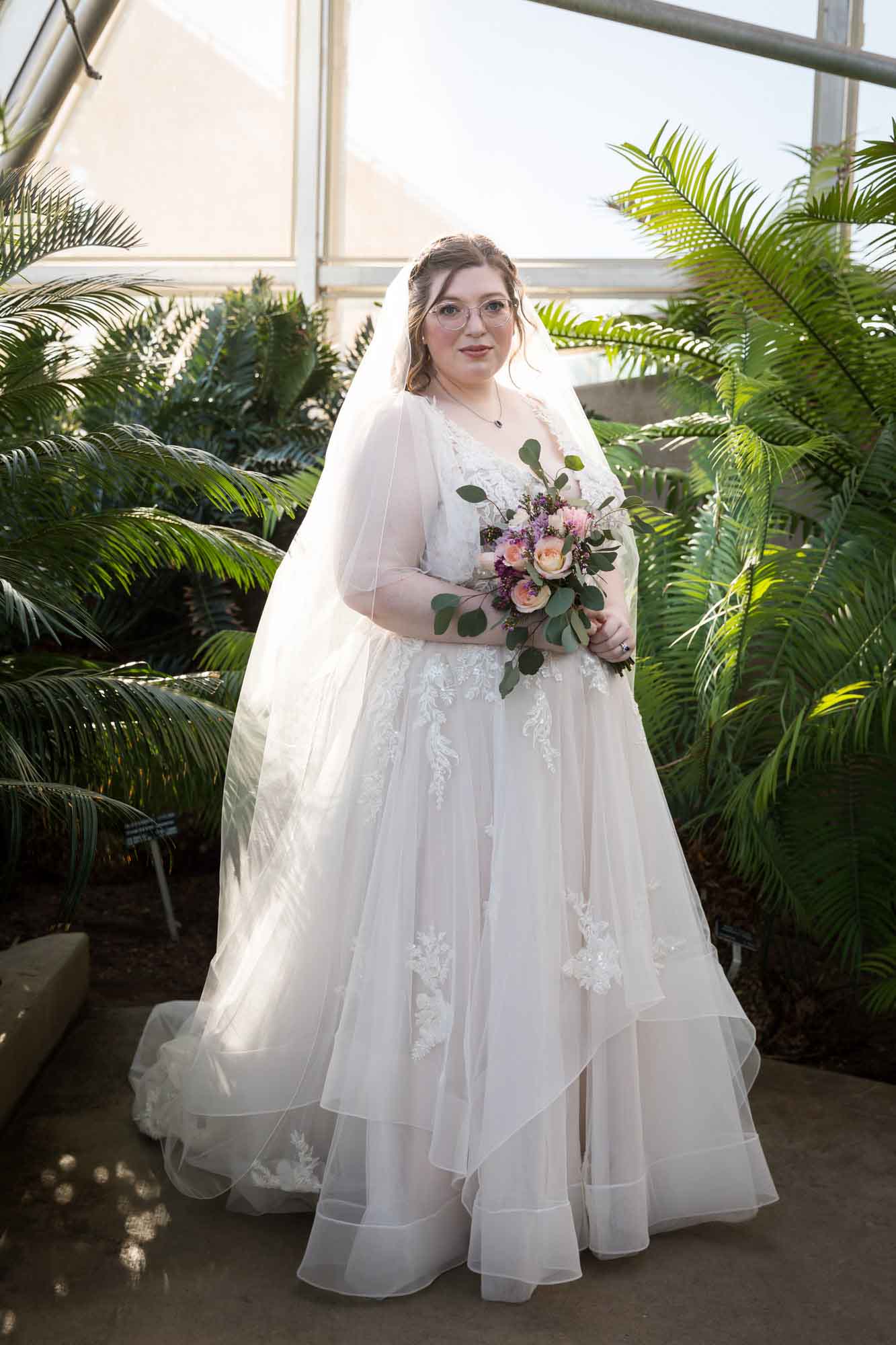 Bride wearing white dress and veil holding flower bouquet standing beside palm branches during a San Antonio Botanical Gardens bridal portrait session