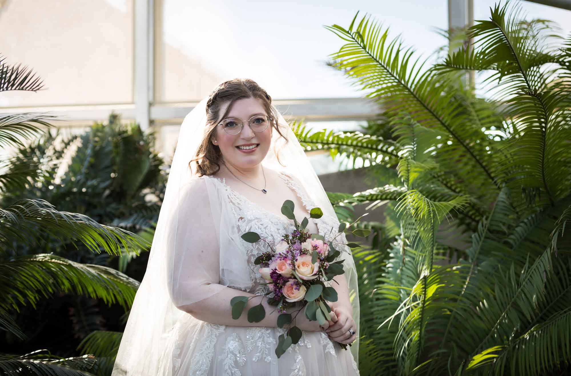 Bride wearing white dress and veil holding flower bouquet standing beside palm branches during a San Antonio Botanical Gardens bridal portrait session