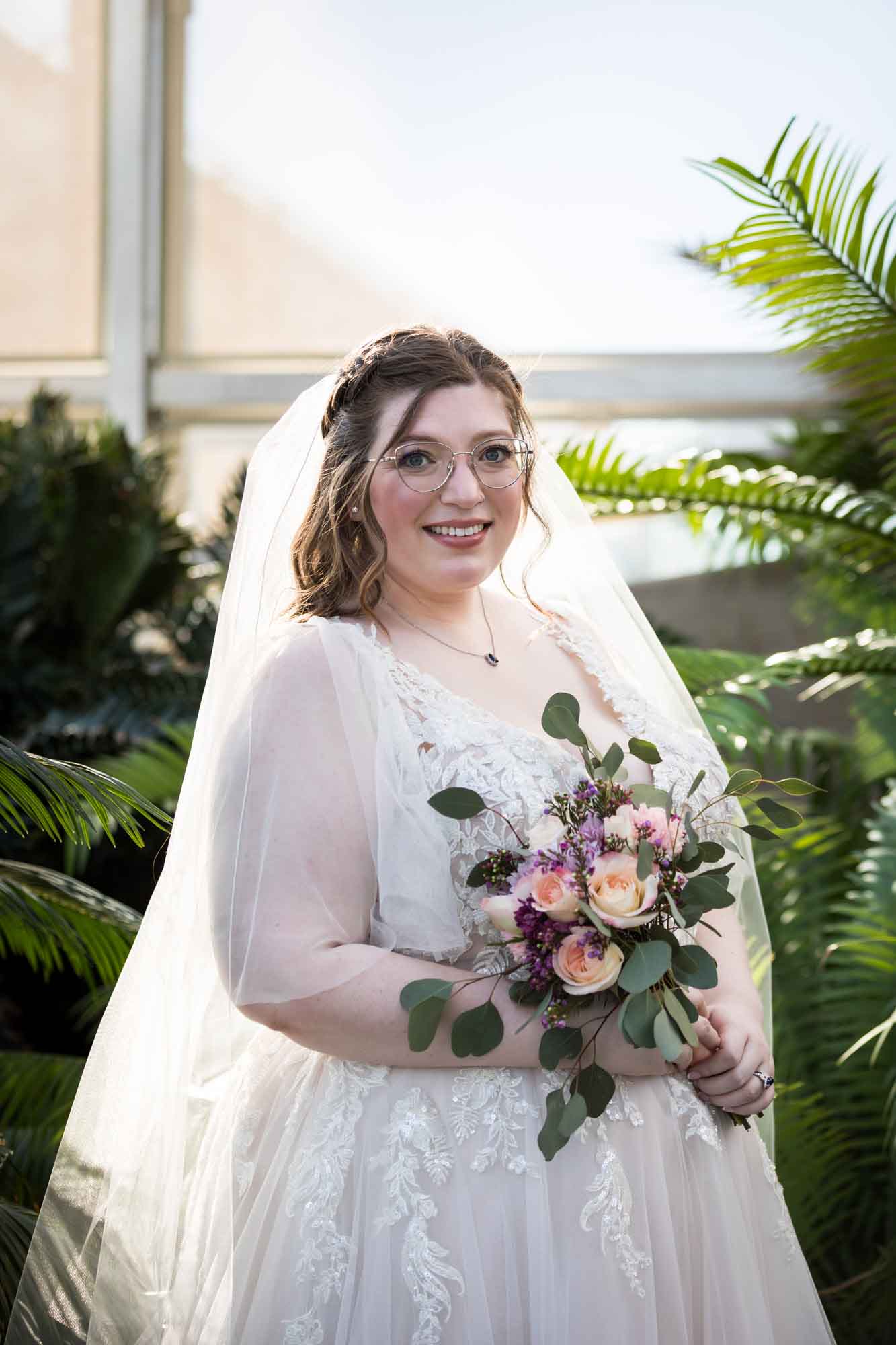 Bride wearing white dress and veil holding flower bouquet standing beside palm branches during a San Antonio Botanical Gardens bridal portrait session