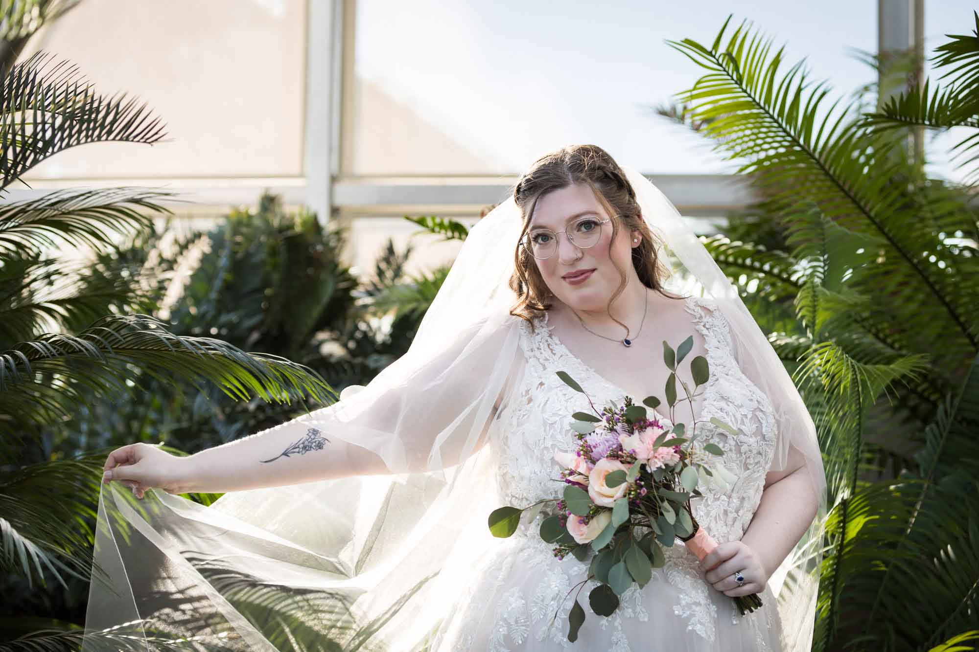 Bride wearing white dress and veil holding flower bouquet and hem of dress standing beside palm branches during a San Antonio Botanical Gardens bridal portrait session