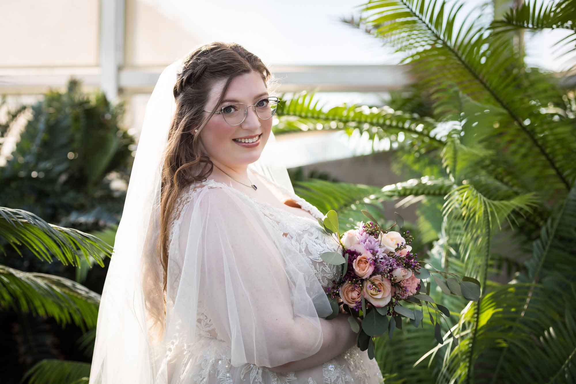 Bride wearing white dress and veil holding flower bouquet standing beside palm branches during a San Antonio Botanical Gardens bridal portrait session