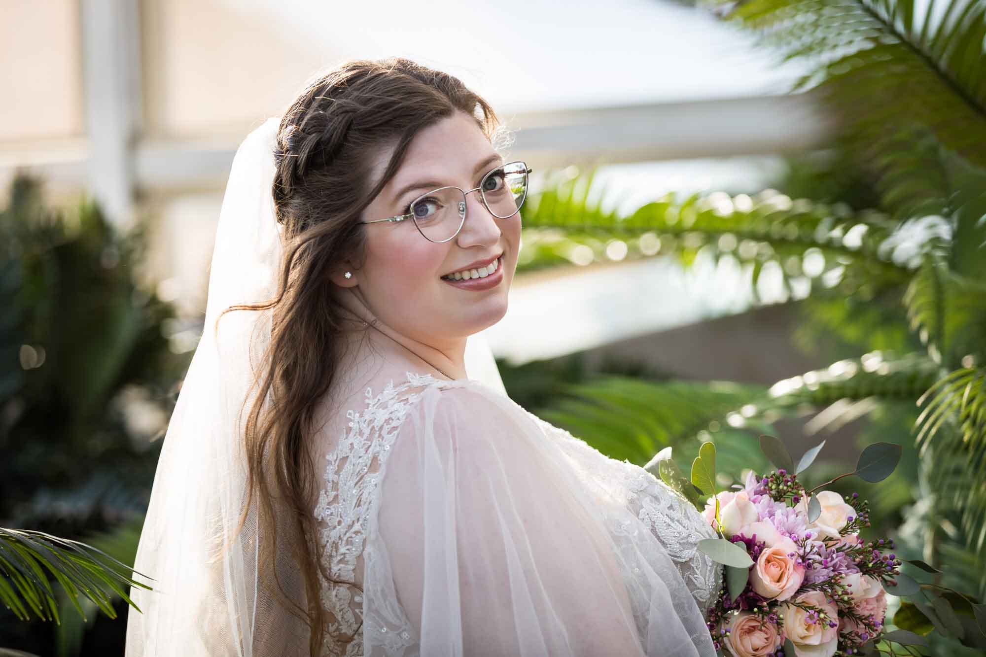 Bride wearing white dress and veil holding flower bouquet standing beside palm branches during a San Antonio Botanical Gardens bridal portrait session