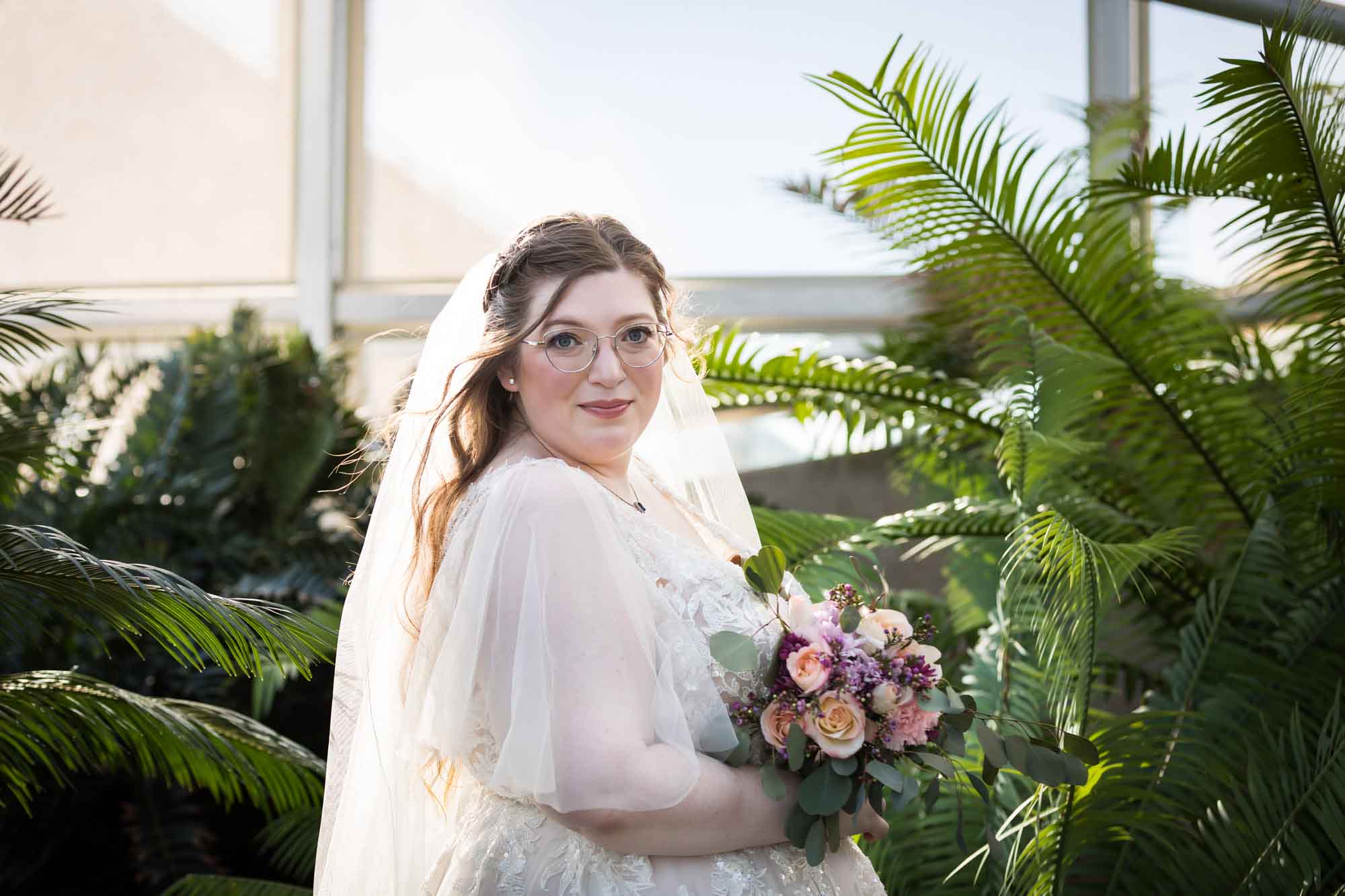 Bride wearing white dress and veil holding flower bouquet standing beside palm branches during a San Antonio Botanical Gardens bridal portrait session