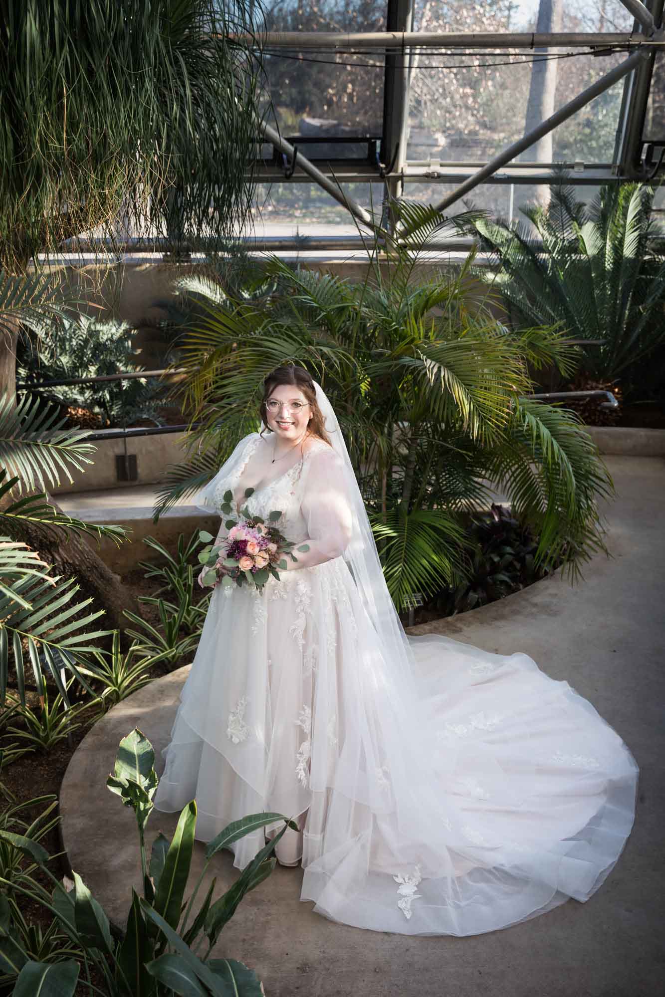 Bride wearing white dress and veil holding flower bouquet surrounded by palms in palm greenhouse during a San Antonio Botanical Gardens bridal portrait session