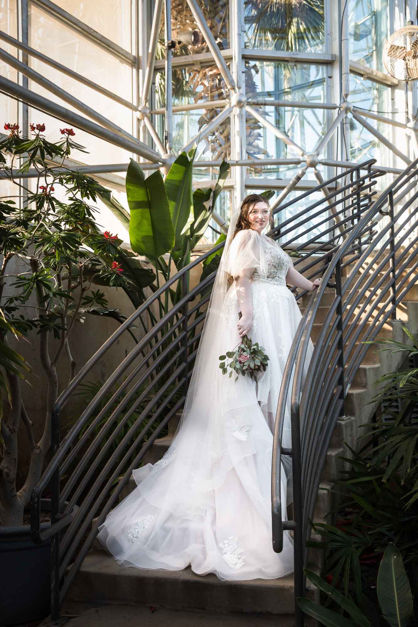 Bride wearing white dress and veil holding flower bouquet and standing on staircase in front of banana tree during a San Antonio Botanical Gardens bridal portrait session