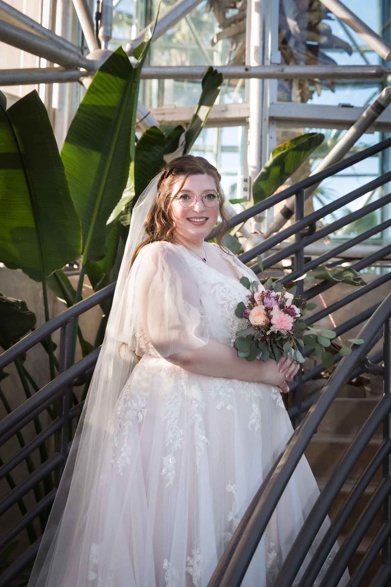 Bride wearing white dress and veil holding flower bouquet and standing on staircase in front of banana tree during a San Antonio Botanical Gardens bridal portrait session