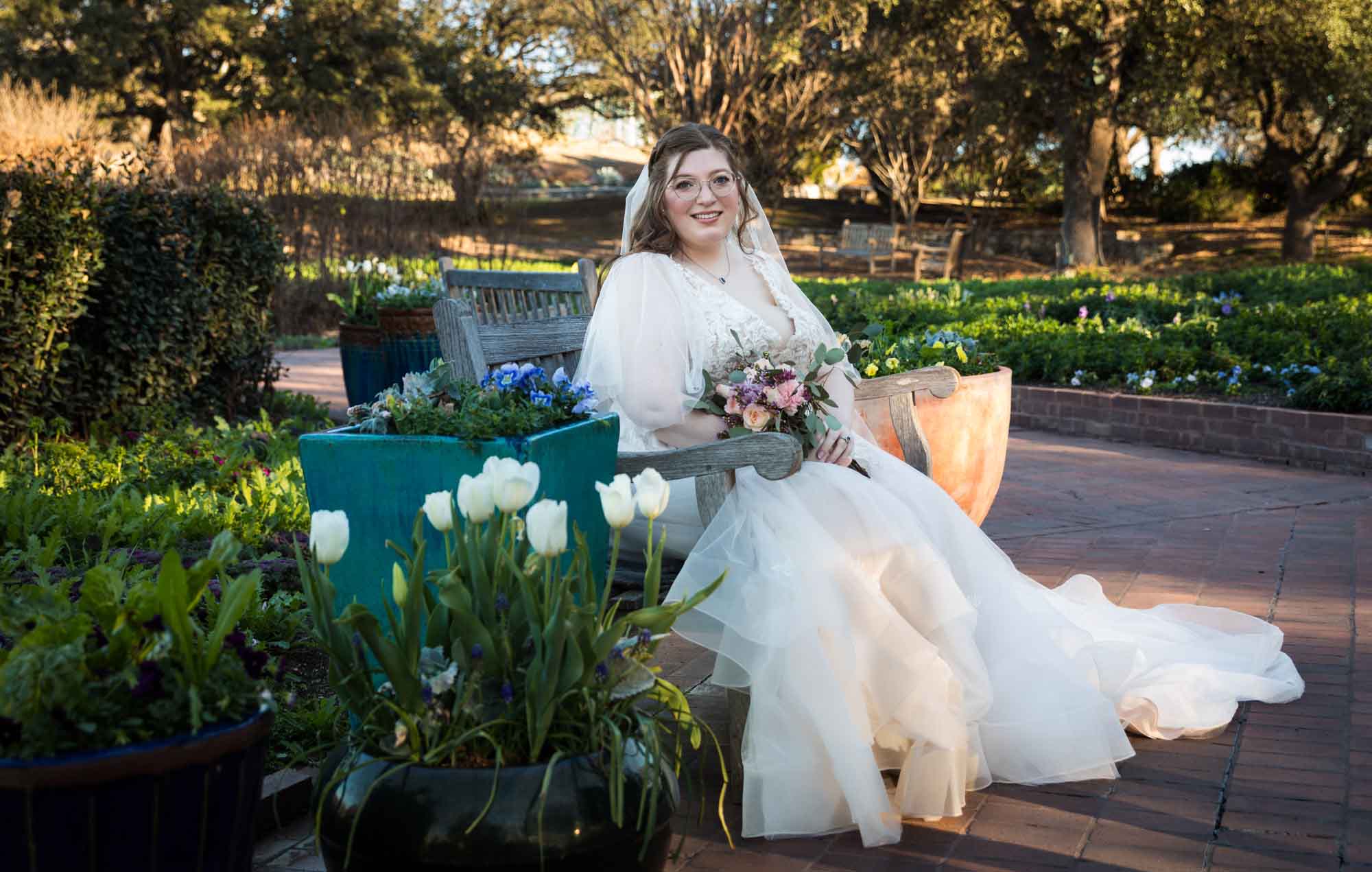 Bride wearing white dress and veil holding flower bouquet and sitting on bench behind colorful flowers during a San Antonio Botanical Gardens bridal portrait session