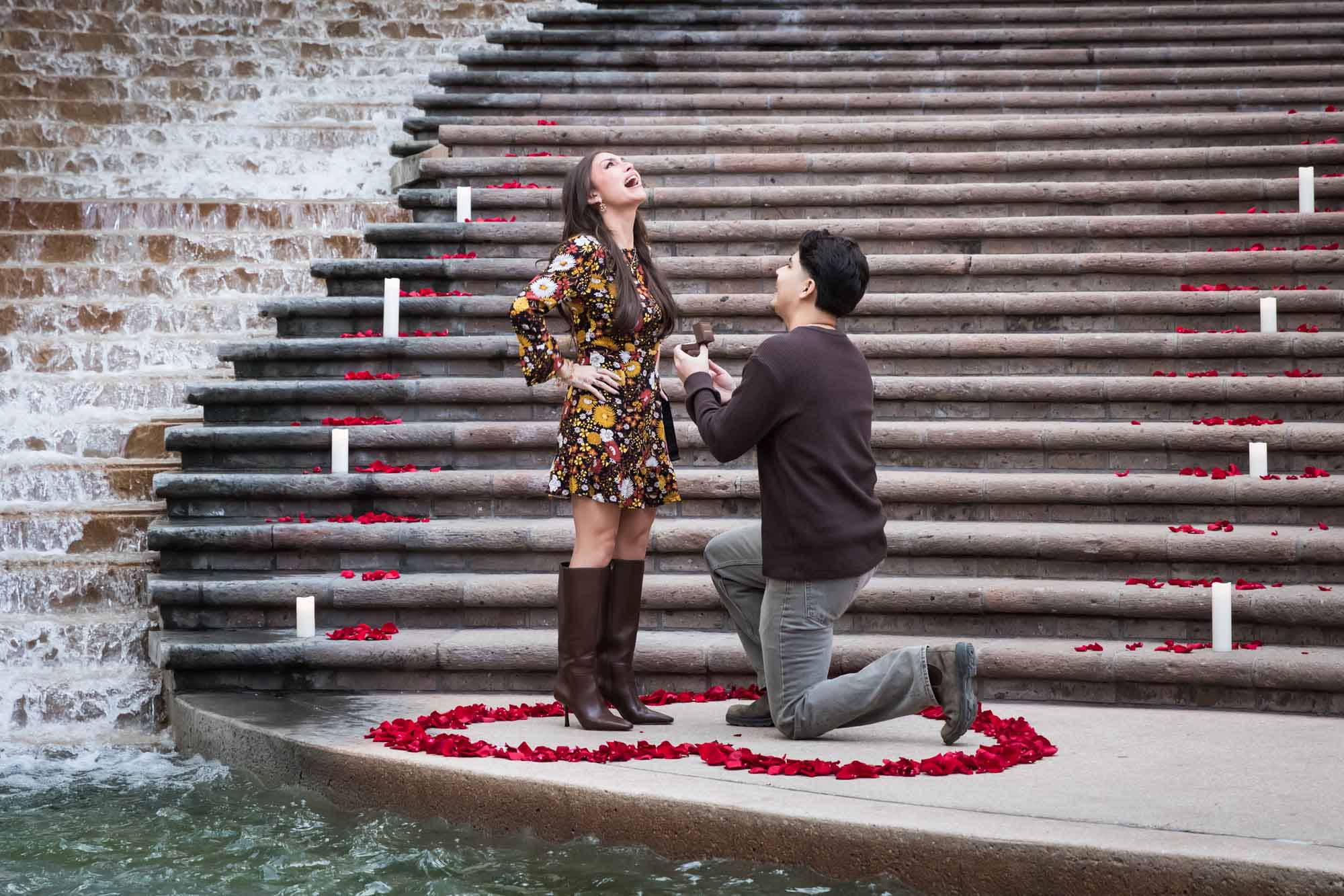 Man proposing to woman on bended knee at the base of the stairs of the Weston Centre in a heart-shaped ring of rose petals during a downtown San Antonio surprise proposal