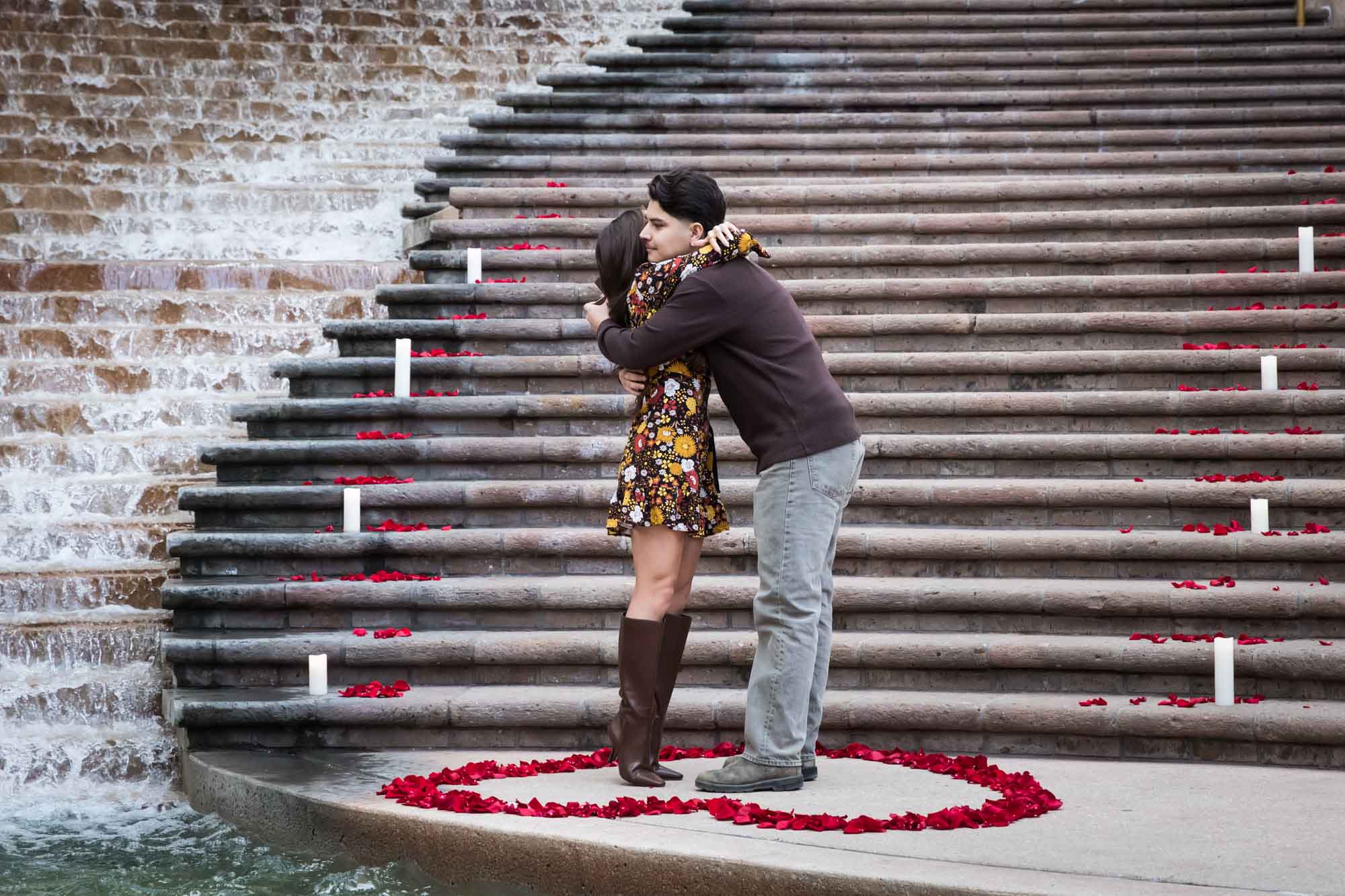 Man hugging woman at the base of the stairs of the Weston Centre in a heart-shaped ring of rose petals during a downtown San Antonio surprise proposal