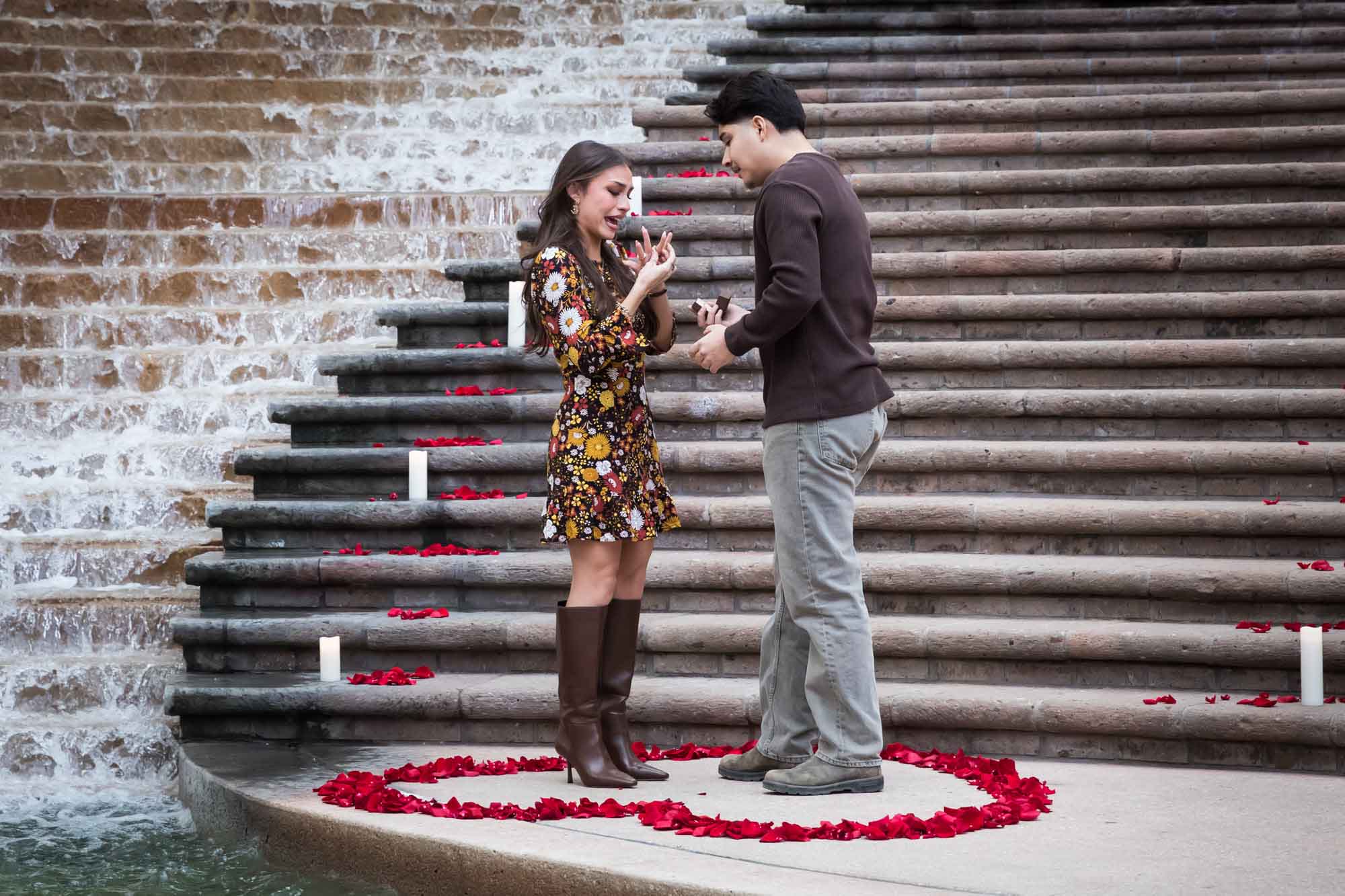 Man proposing to woman at the base of the stairs of the Weston Centre in a heart-shaped ring of rose petals during a downtown San Antonio surprise proposal