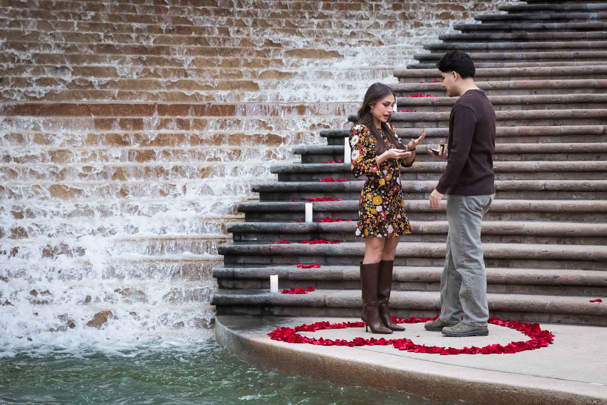 Man proposing to woman at the base of the stairs of the Weston Centre in a heart-shaped ring of rose petals during a downtown San Antonio surprise proposal