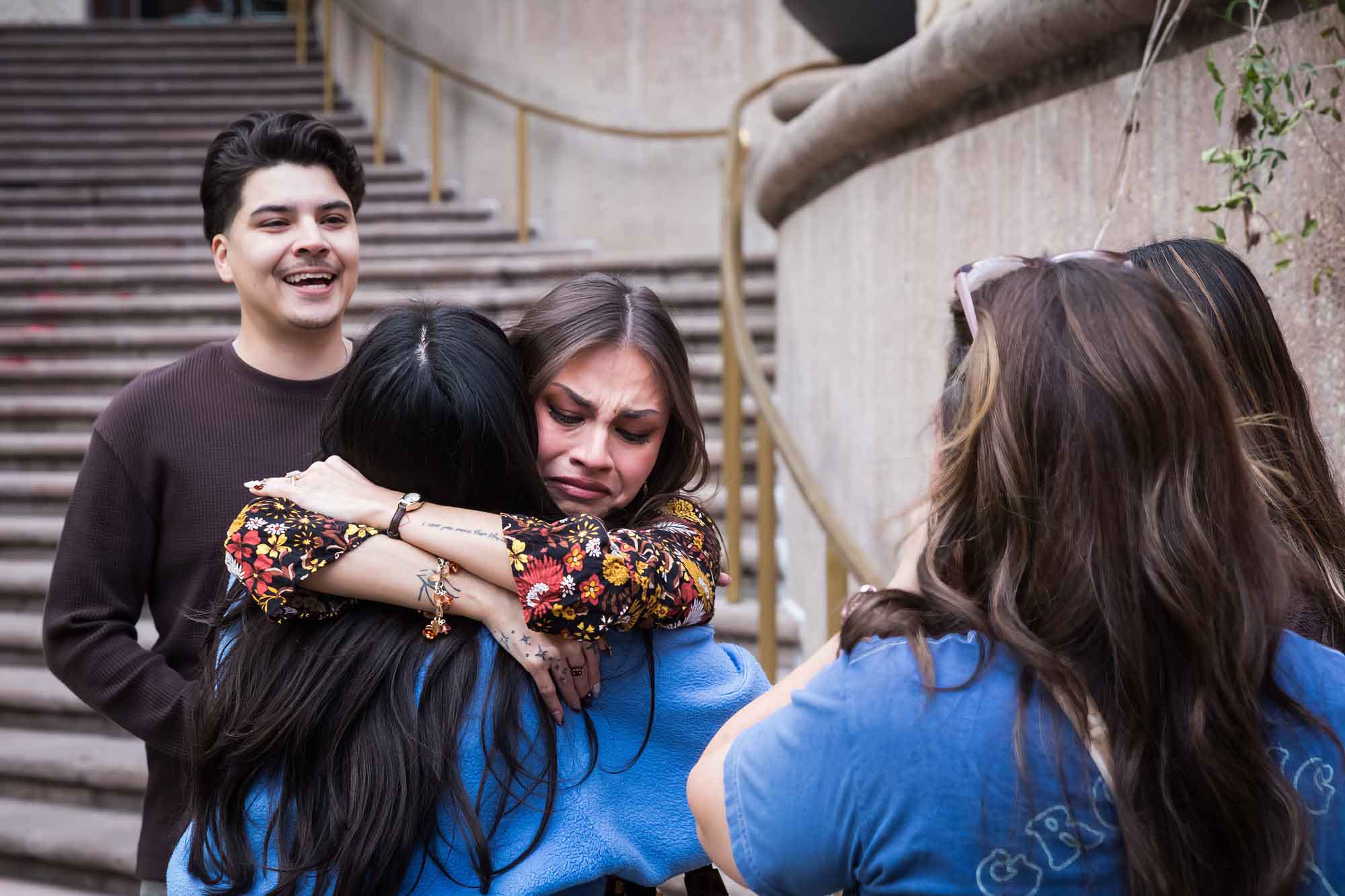 Two women hugging in front of man and two other women in front of stairs during a downtown San Antonio surprise proposal