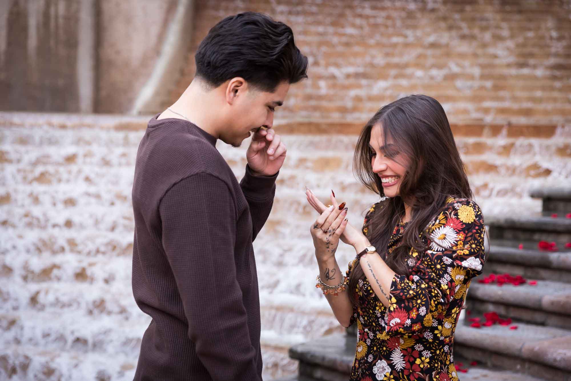 Man and woman looking at engagement ring on woman's finger in front of waterfall during a downtown San Antonio surprise proposal