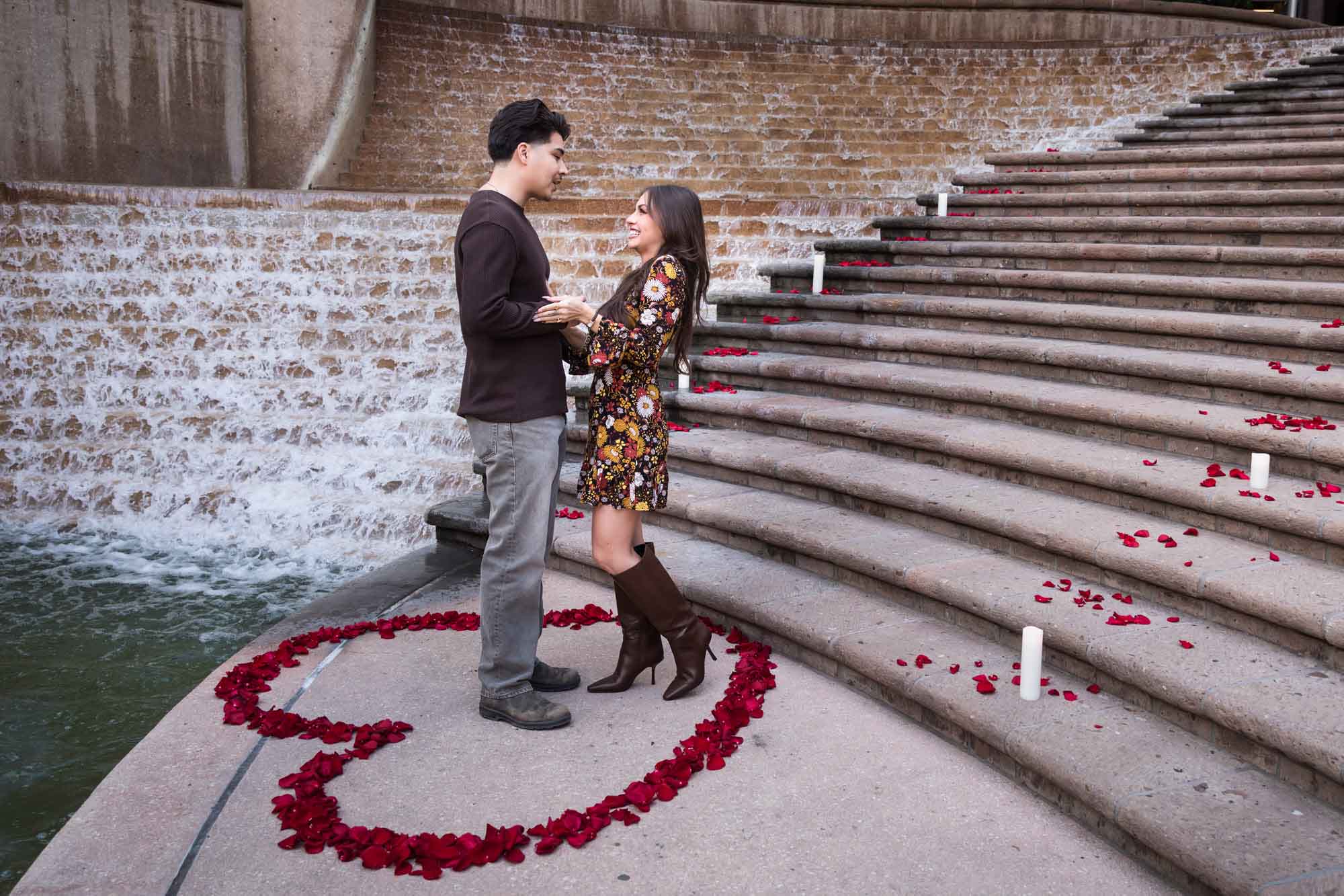 Couple standing in middle of heart-shaped ring of rose petals at bottom of stairs during a downtown San Antonio surprise proposal