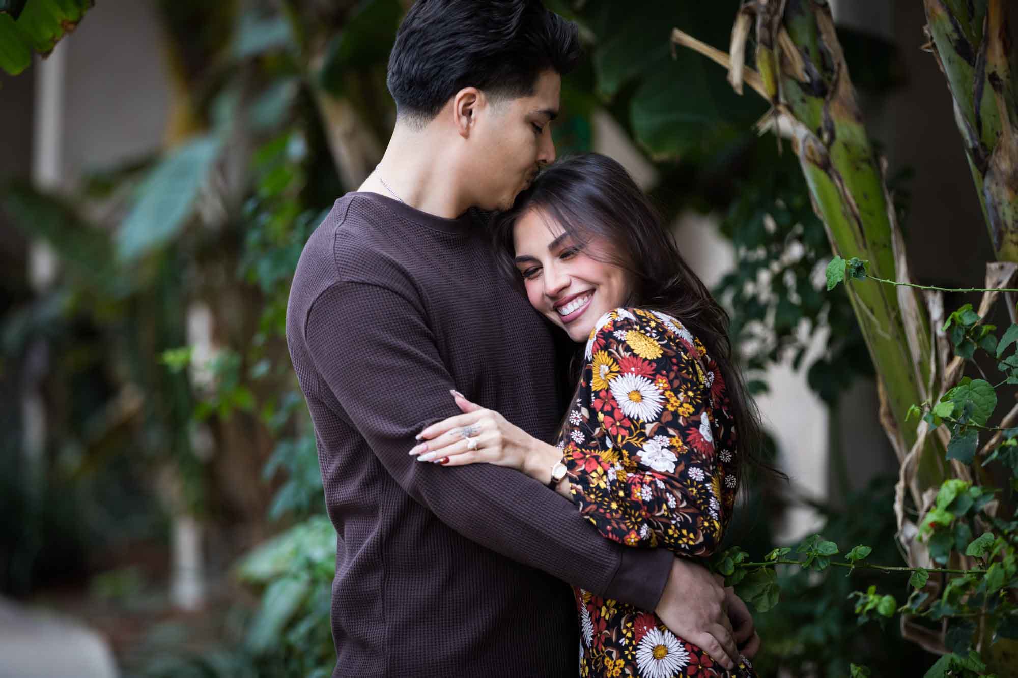 Man kissing woman on head in lush landscaping of River Walk during a downtown San Antonio surprise proposal