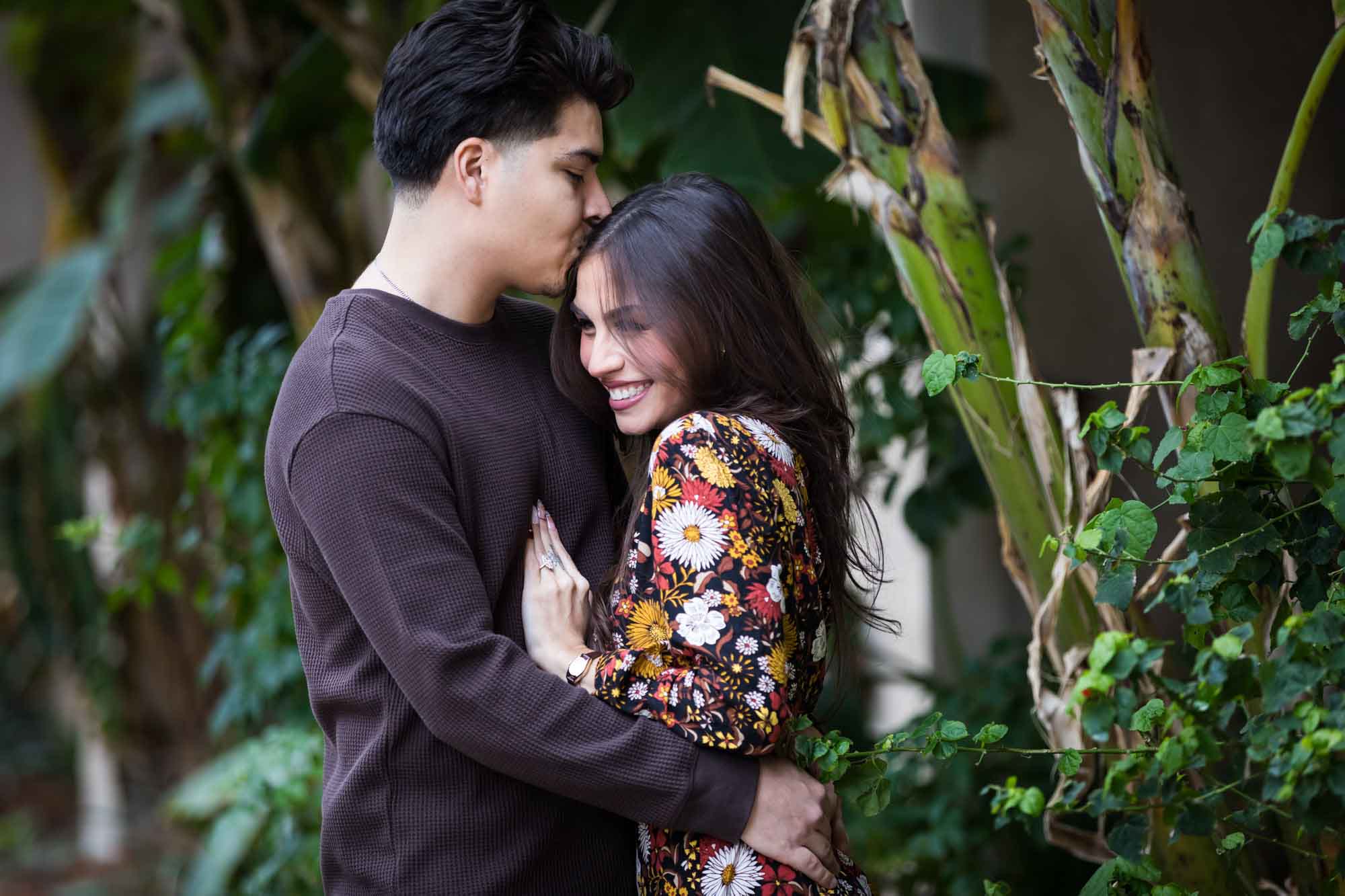 Man kissing woman on head in lush landscaping of River Walk during a downtown San Antonio surprise proposal