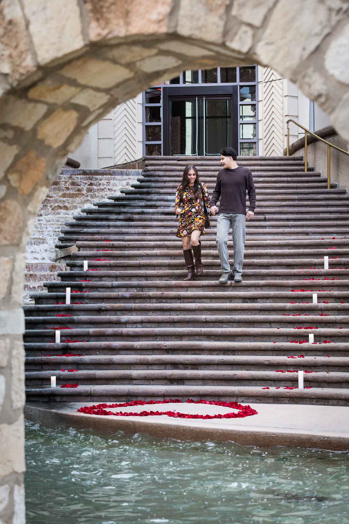 Couple walking down the stairs of the Weston Centre to a heart-shaped ring of rose petals during a downtown San Antonio surprise proposal