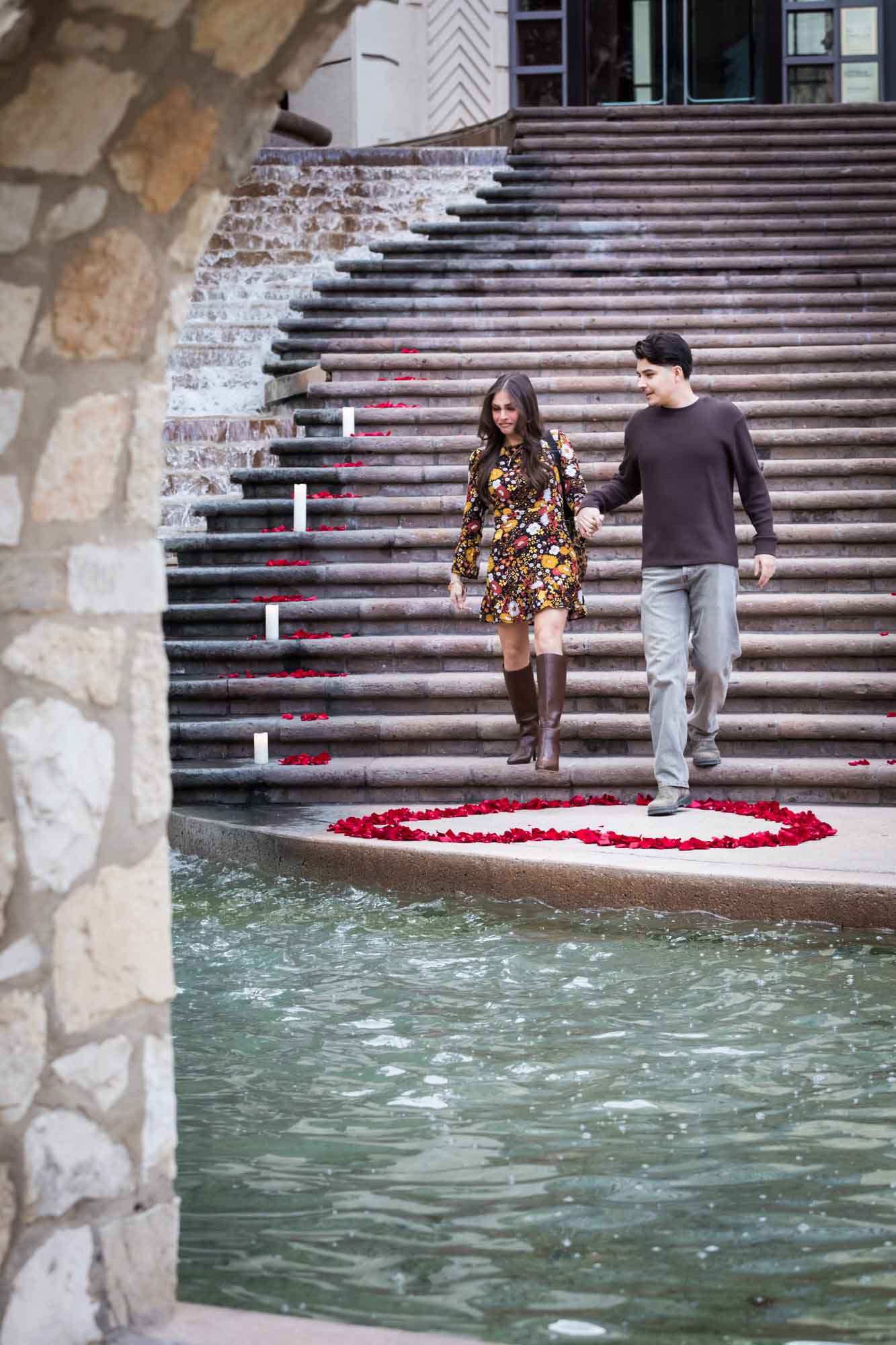 Couple walking down the stairs of the Weston Centre to a heart-shaped ring of rose petals during a downtown San Antonio surprise proposal