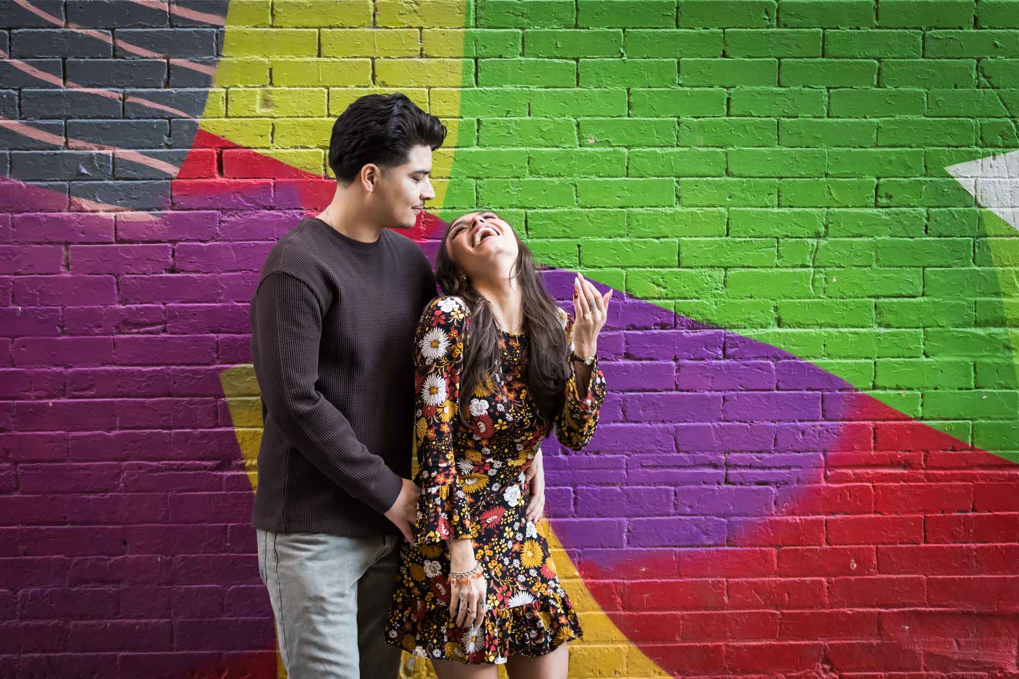 Woman laughing in front of man in front of colorful brick wall during a downtown San Antonio surprise proposal