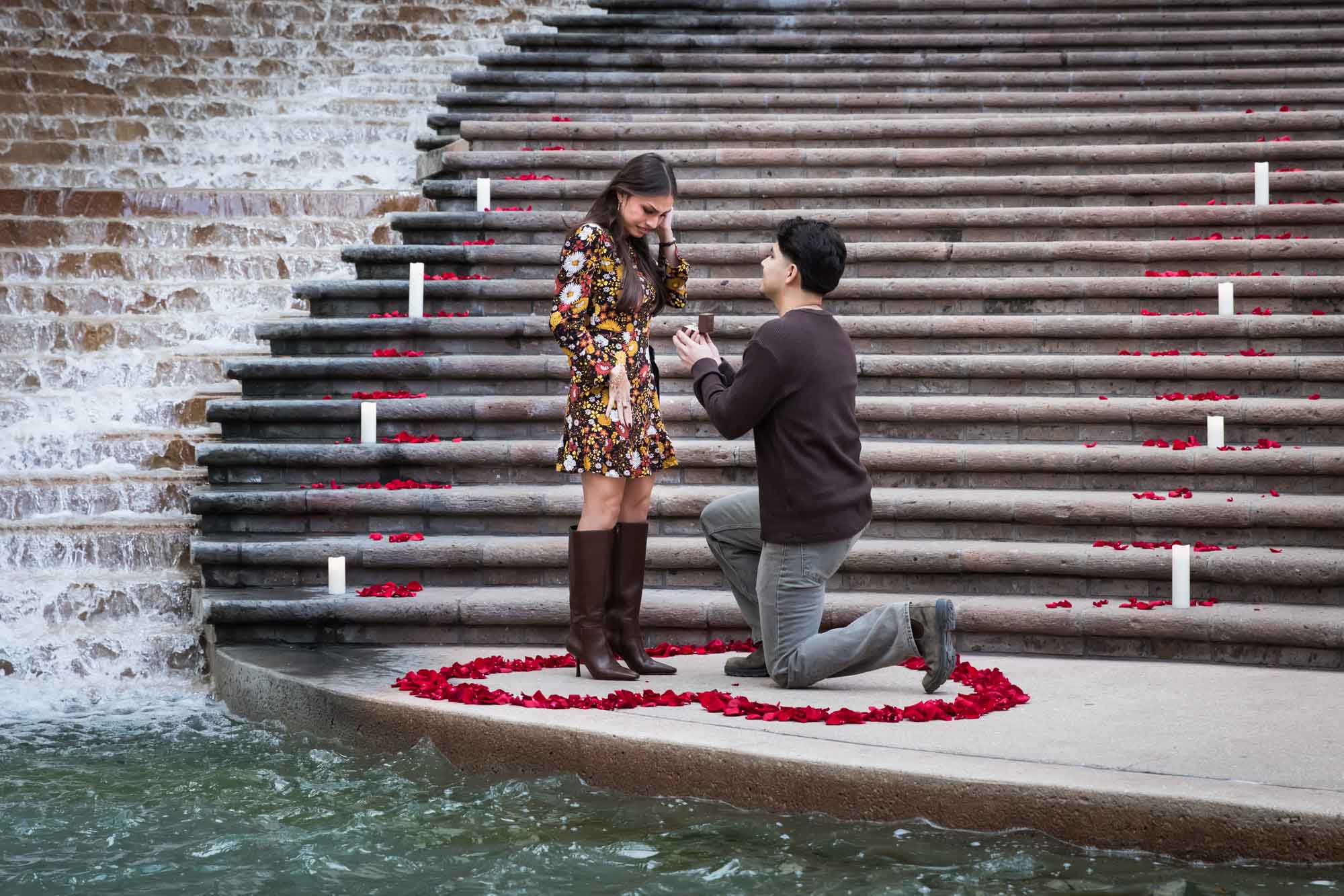 Man proposing to woman on bended knee at the base of the stairs of the Weston Centre in a heart-shaped ring of rose petals during a downtown San Antonio surprise proposal