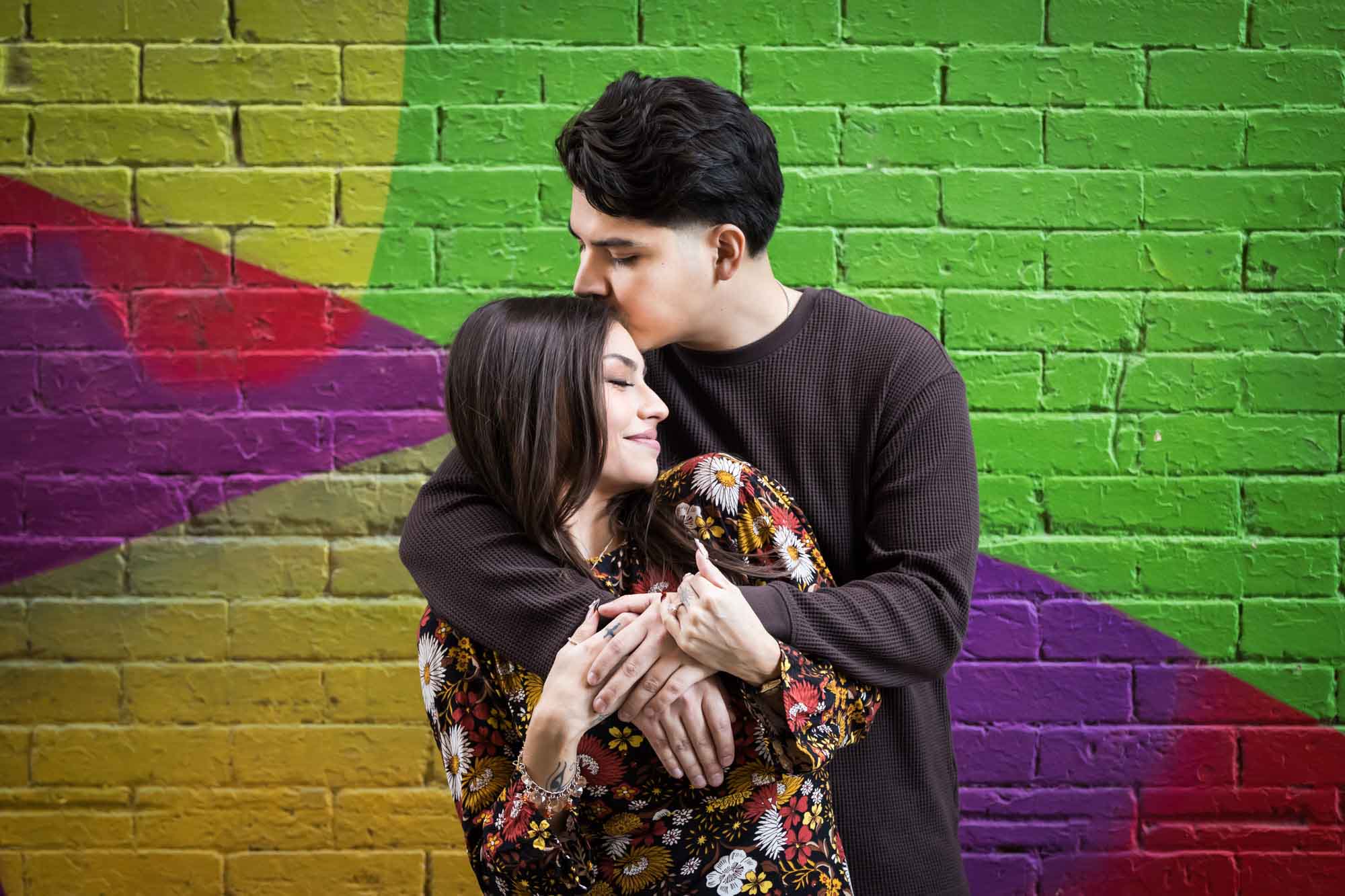Man kissing woman's forehead in front of colorful brick wall during a downtown San Antonio surprise proposal